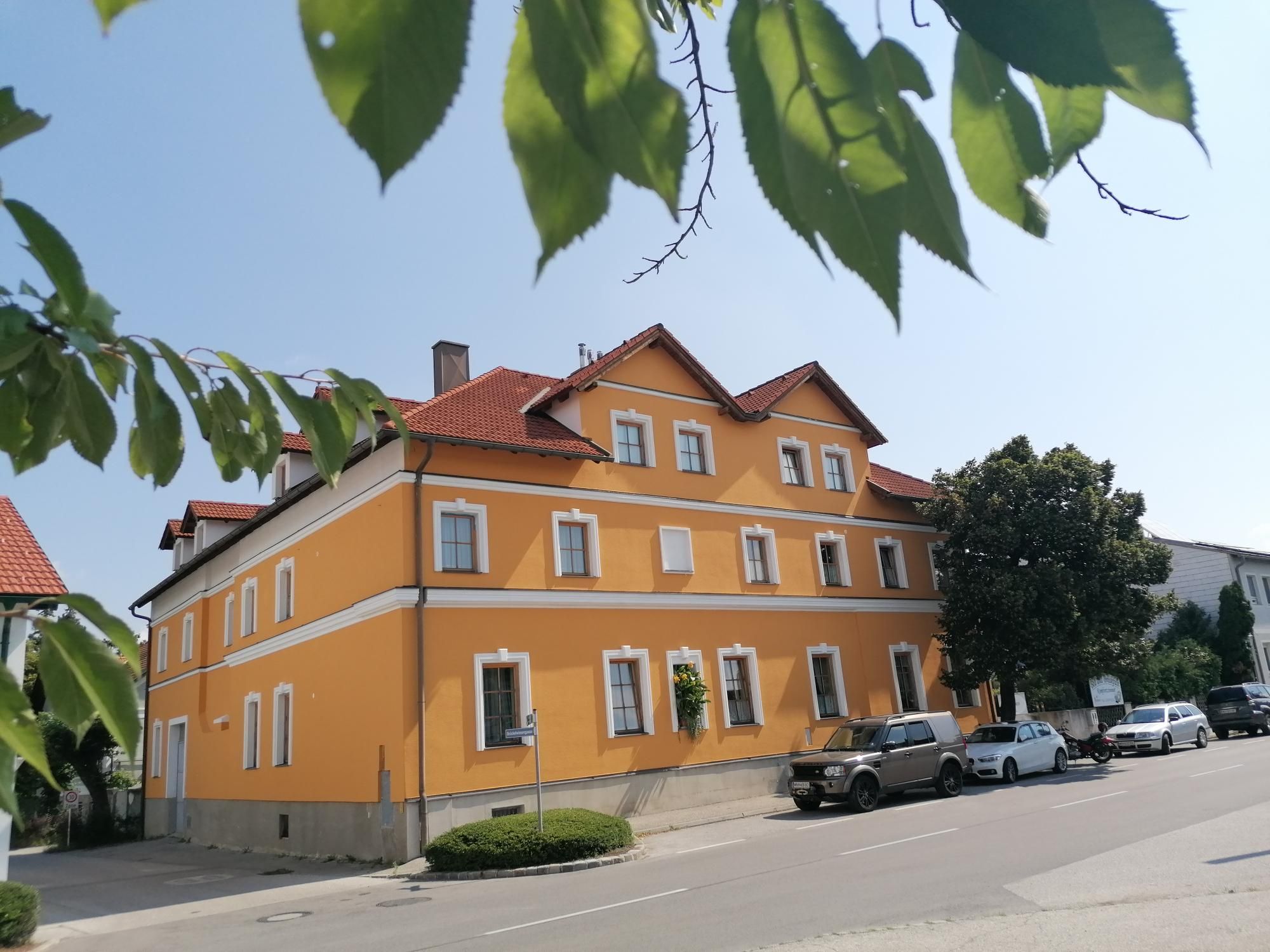 Sárga épület piros tetővel, fákkal és parkoló autókkal körülvéve.