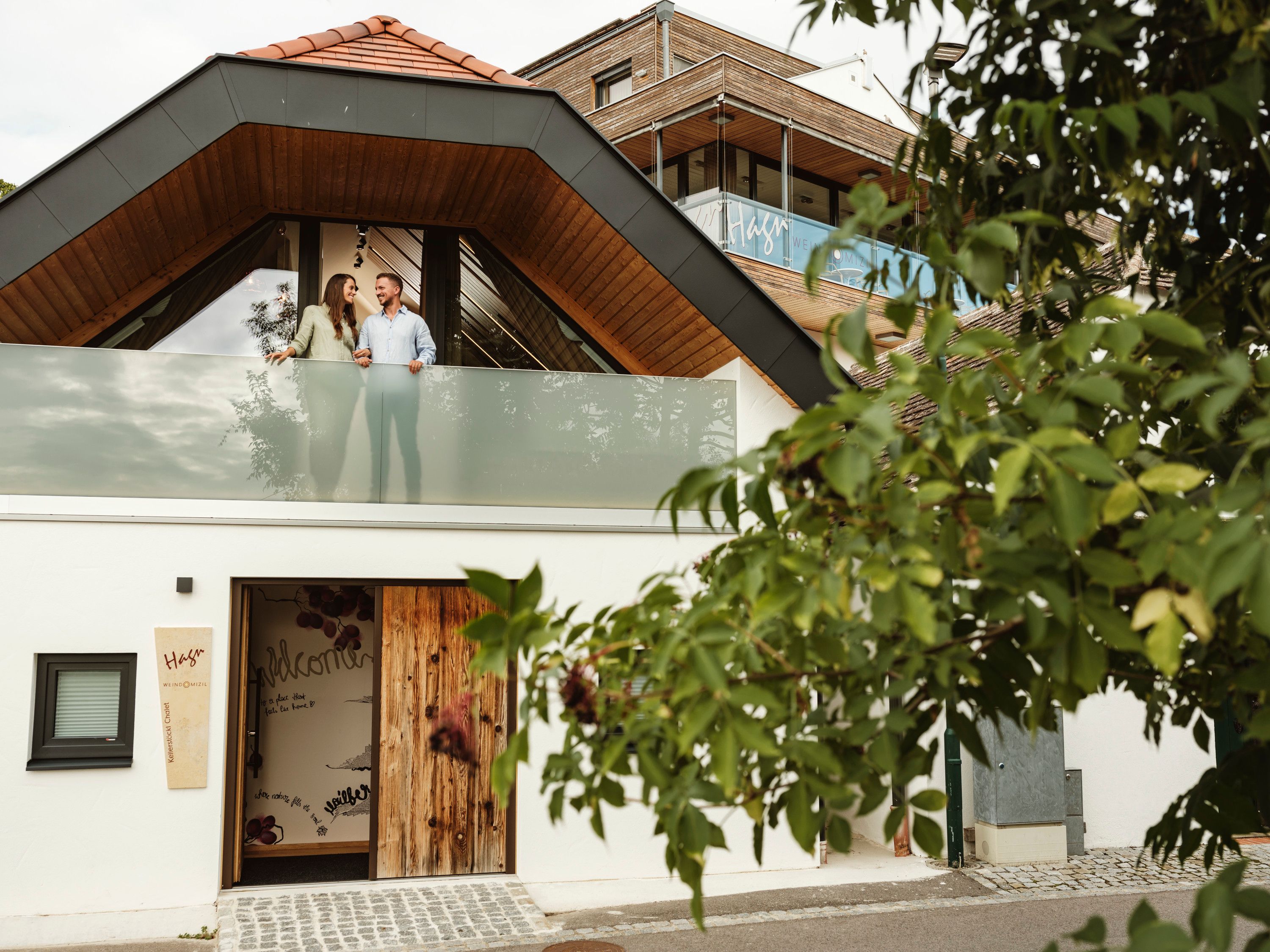 Modern épület, párral az erkélyen, fákkal körülvéve.