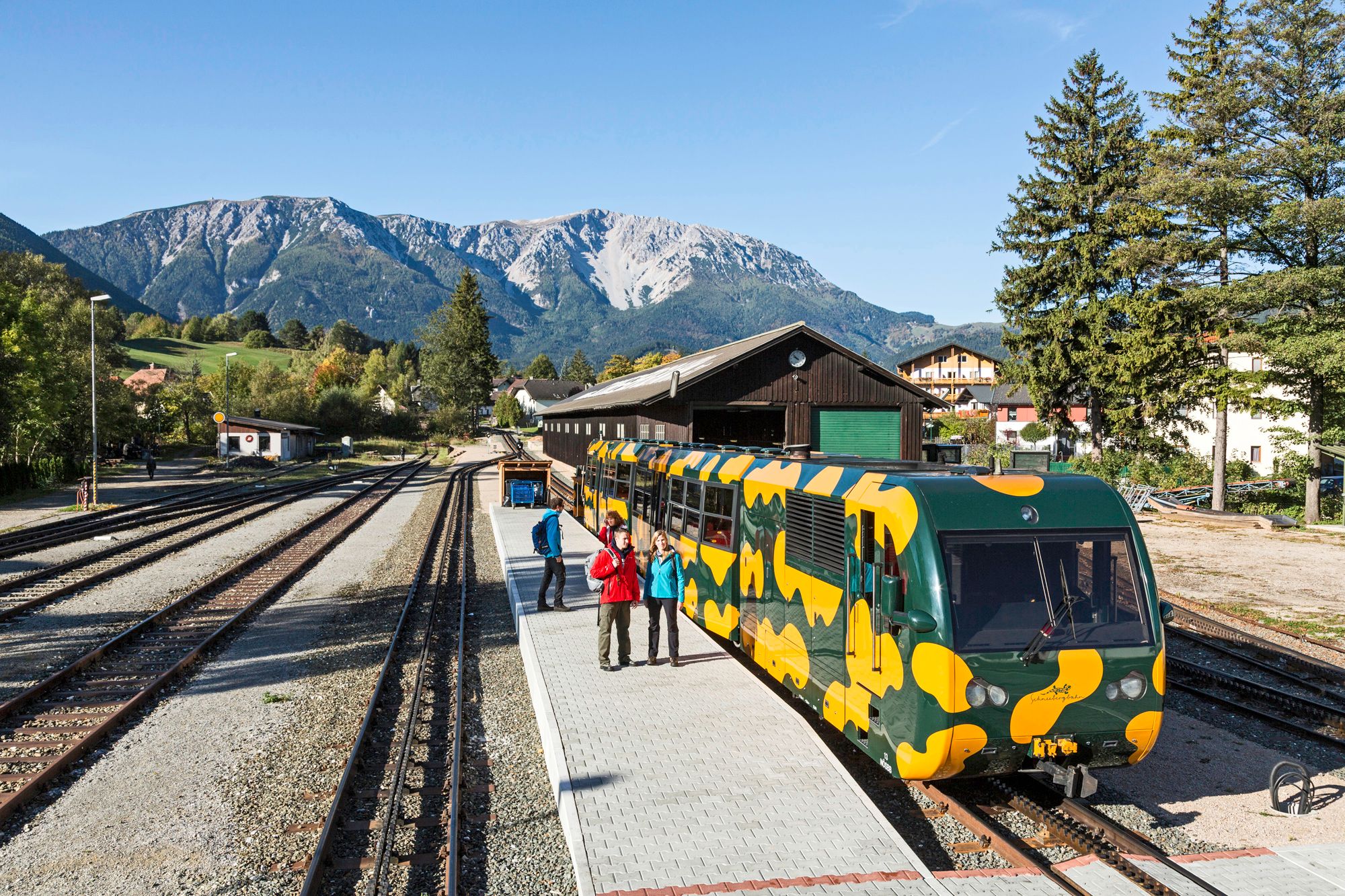 Puchberg am Schneeberg vasútállomás, előtérben színes fogaskerekű vasút, háttérben hegyek.