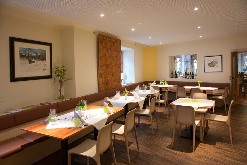 Modern étkező terített asztalokkal, székekkel és dekorációval. A falakon képek lógnak, és van egy ablakpárkány borosüvegekkel.