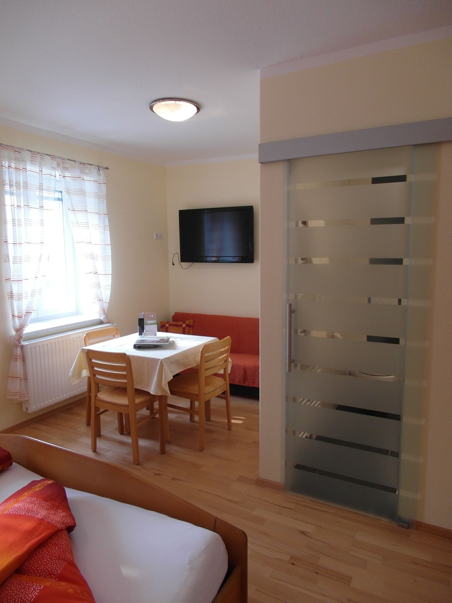 Világos szoba étkezővel, asztallal, székekkel, ággyal és síkképernyős TV-vel.