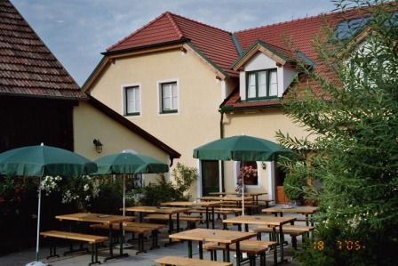 Egy borozó kertje fapadokkal és zöld napernyőkkel egy sárga, piros tetőcseréppel fedett épület előtt.