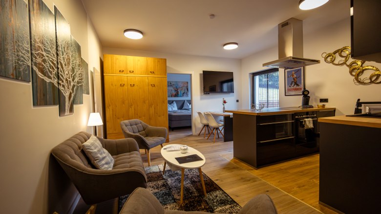 Modern nappali konyhával, kanapéval, fotelekkel és étkezőasztallal. Világos színű fabútorok és faliképek láthatók.