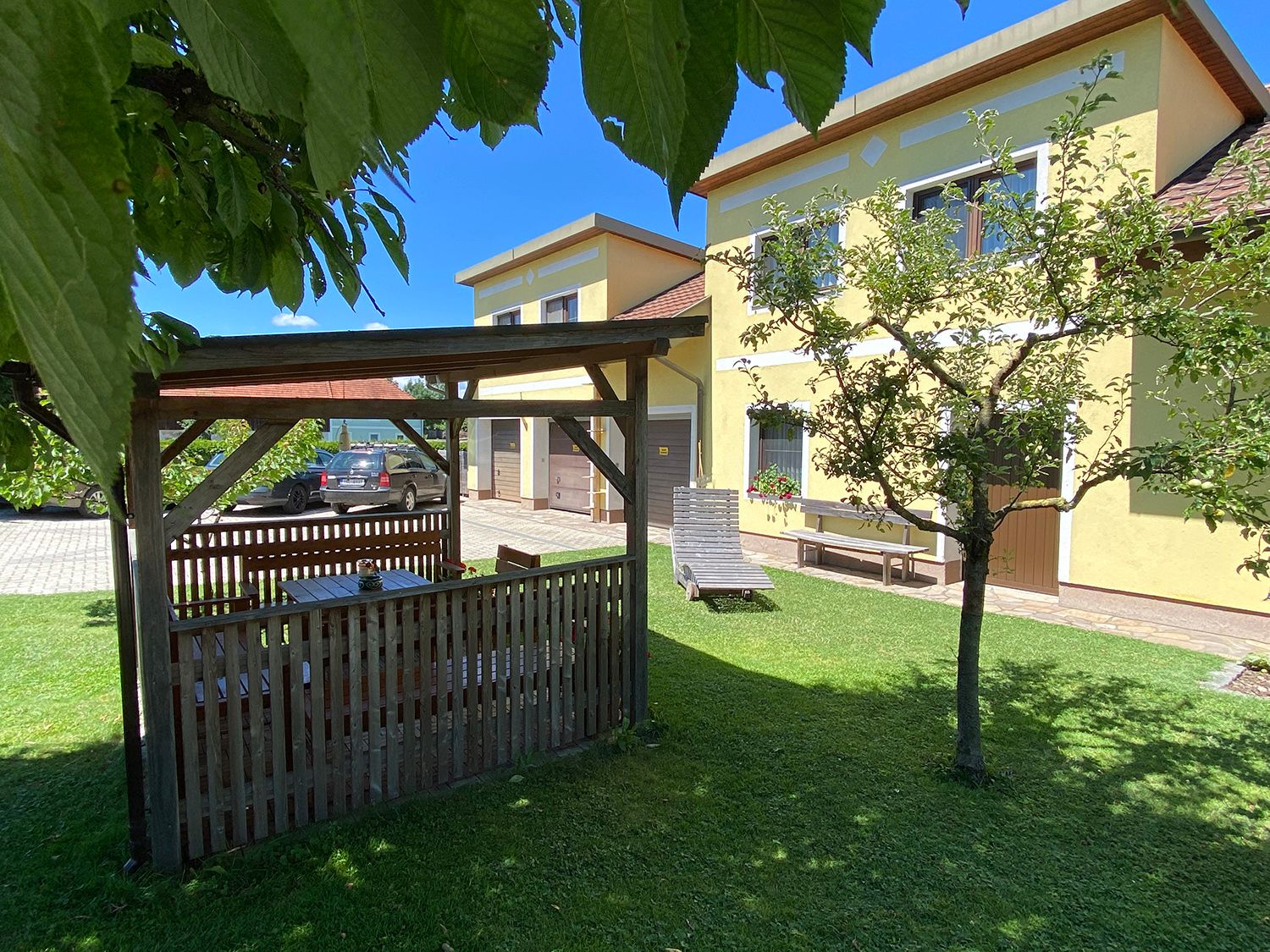 Kert pavilonnal, sárga házzal és autóval a háttérben.