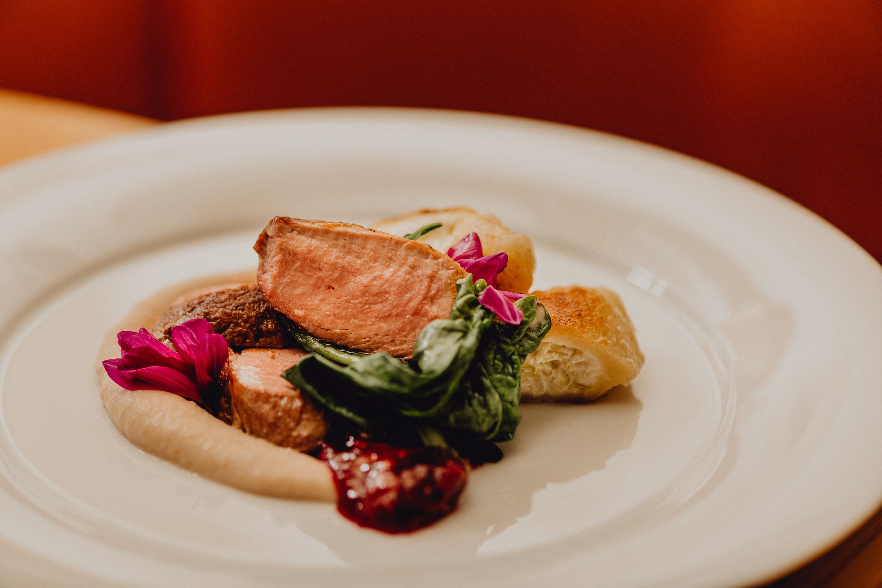 Egy tányér rózsaszínű sült kacsa, tejszínes lepény, gesztenyepüré és zöld levelek, ehető virágokkal díszítve.