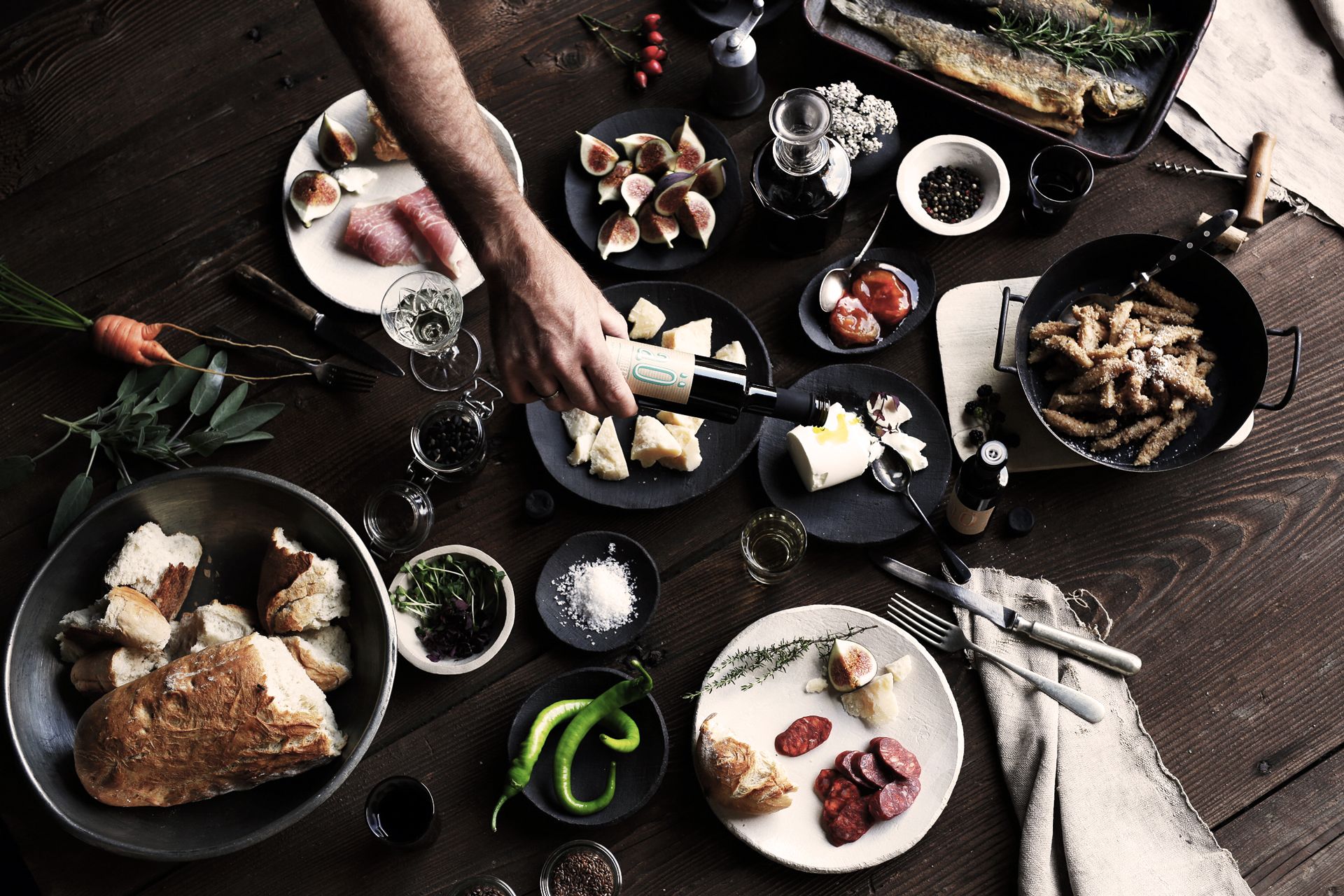 Egy rusztikus asztal különböző ételekkel, többek között kenyérrel, sajttal, fügével, kolbásszal és hallal. Egy kéz olajat önt az egyik tálra.