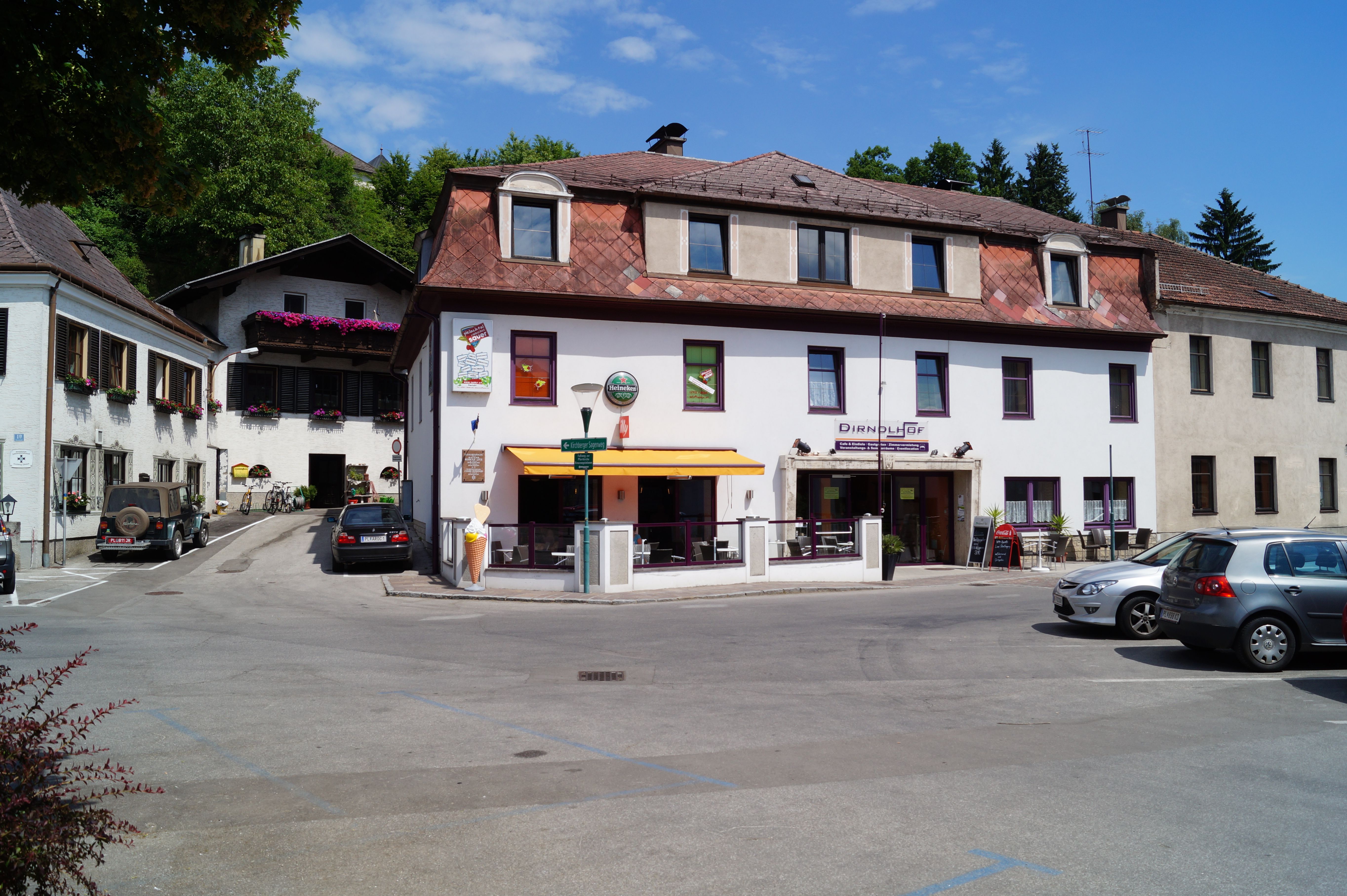Egy "Dirndlhof" feliratú épület és egy napernyős terasz. Az autók az előtte lévő parkolóban parkolnak.