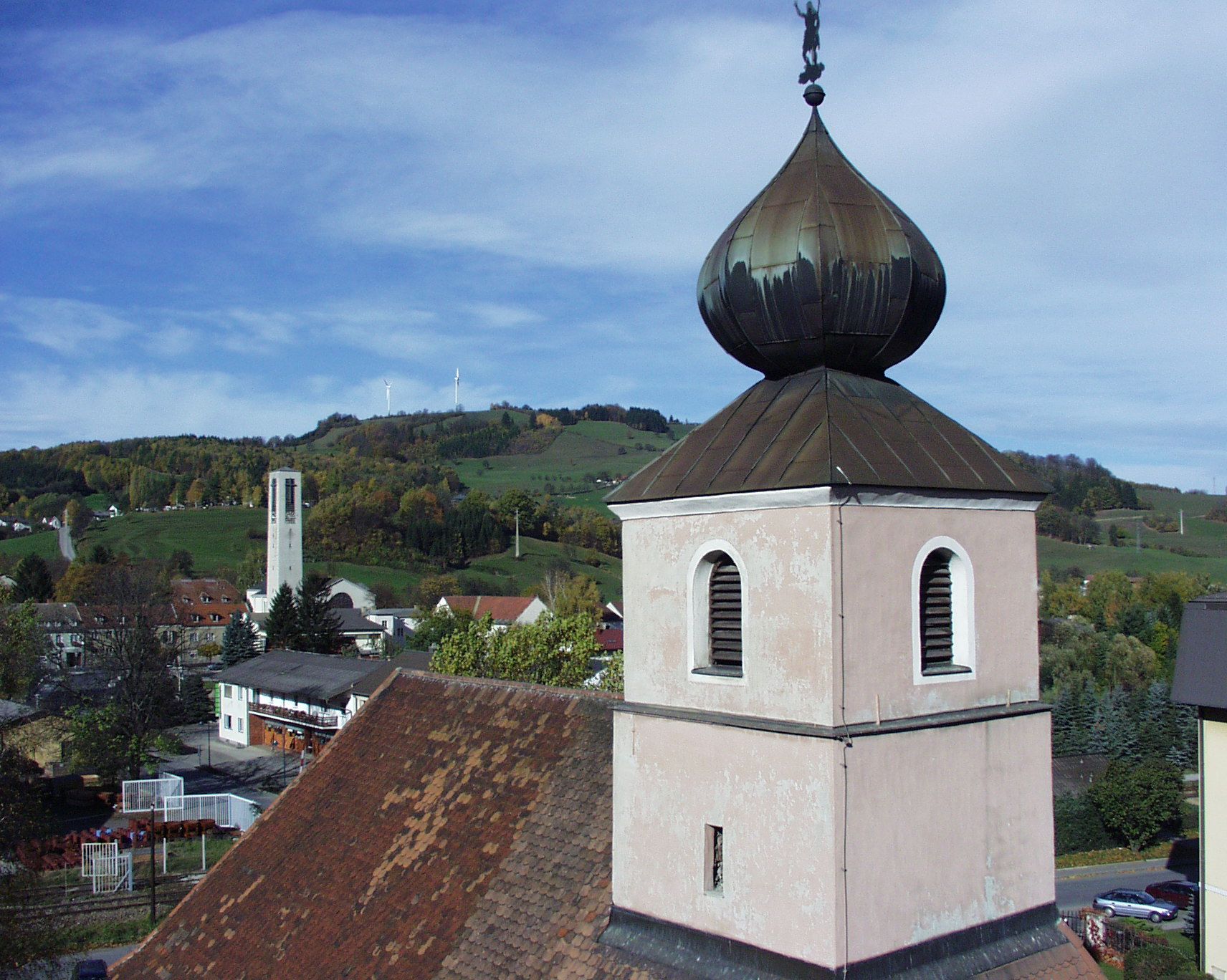 Hagymakupolás templomtorony dombos táj és szélturbinák előtt.