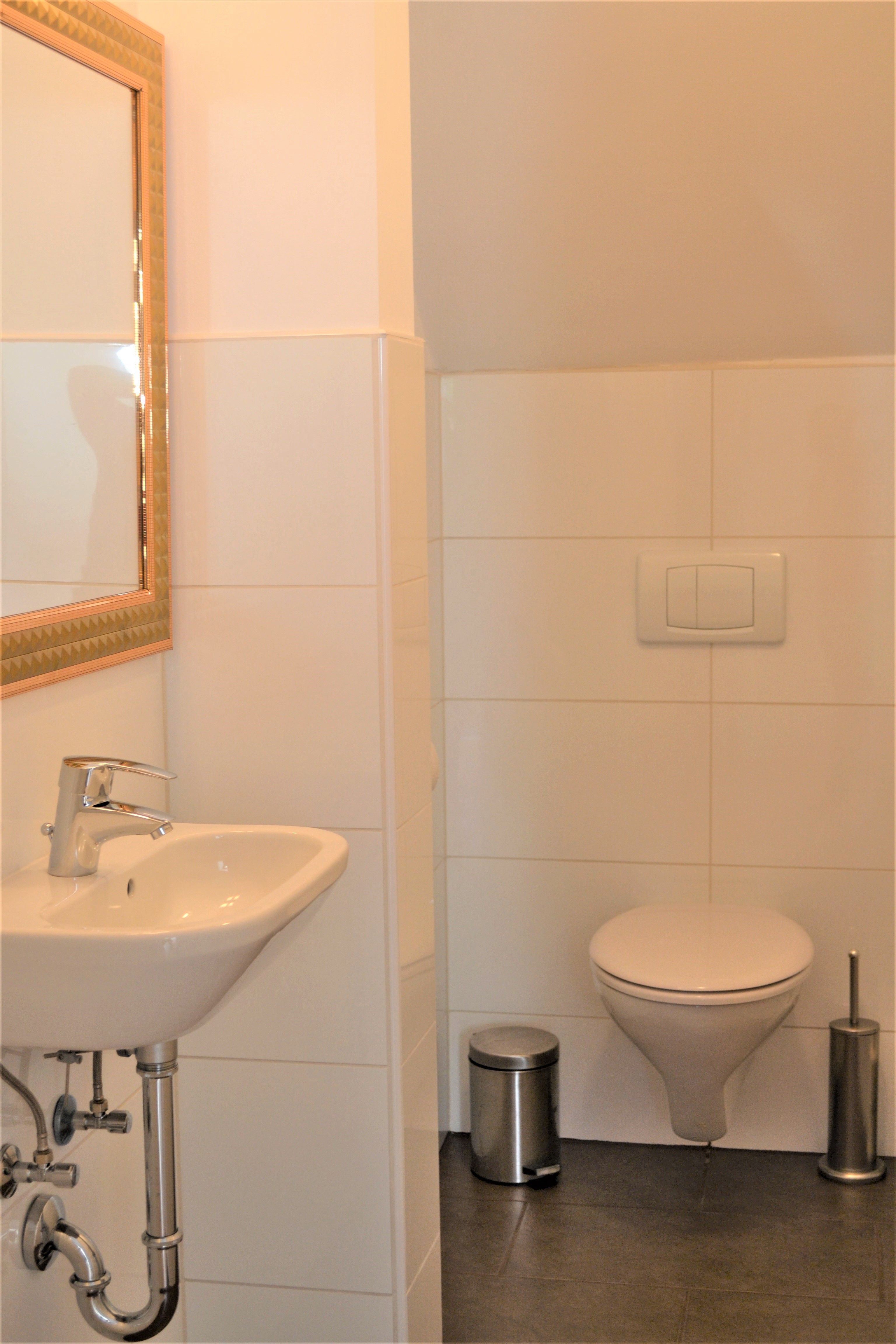 Kis fürdőszoba mosdóval, tükörrel, WC-vel, szemetesládával és WC-kefével.