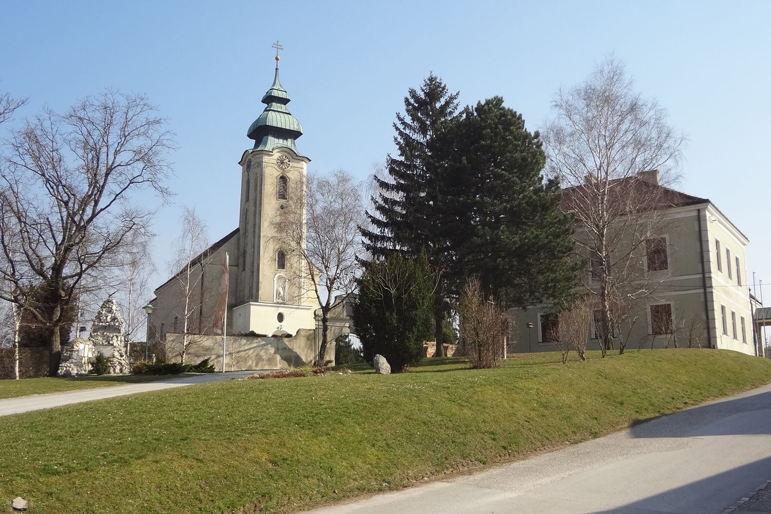 Templom toronnyal és szomszédos épülettel Gnadendorfban, fákkal és pázsittal körülvéve.