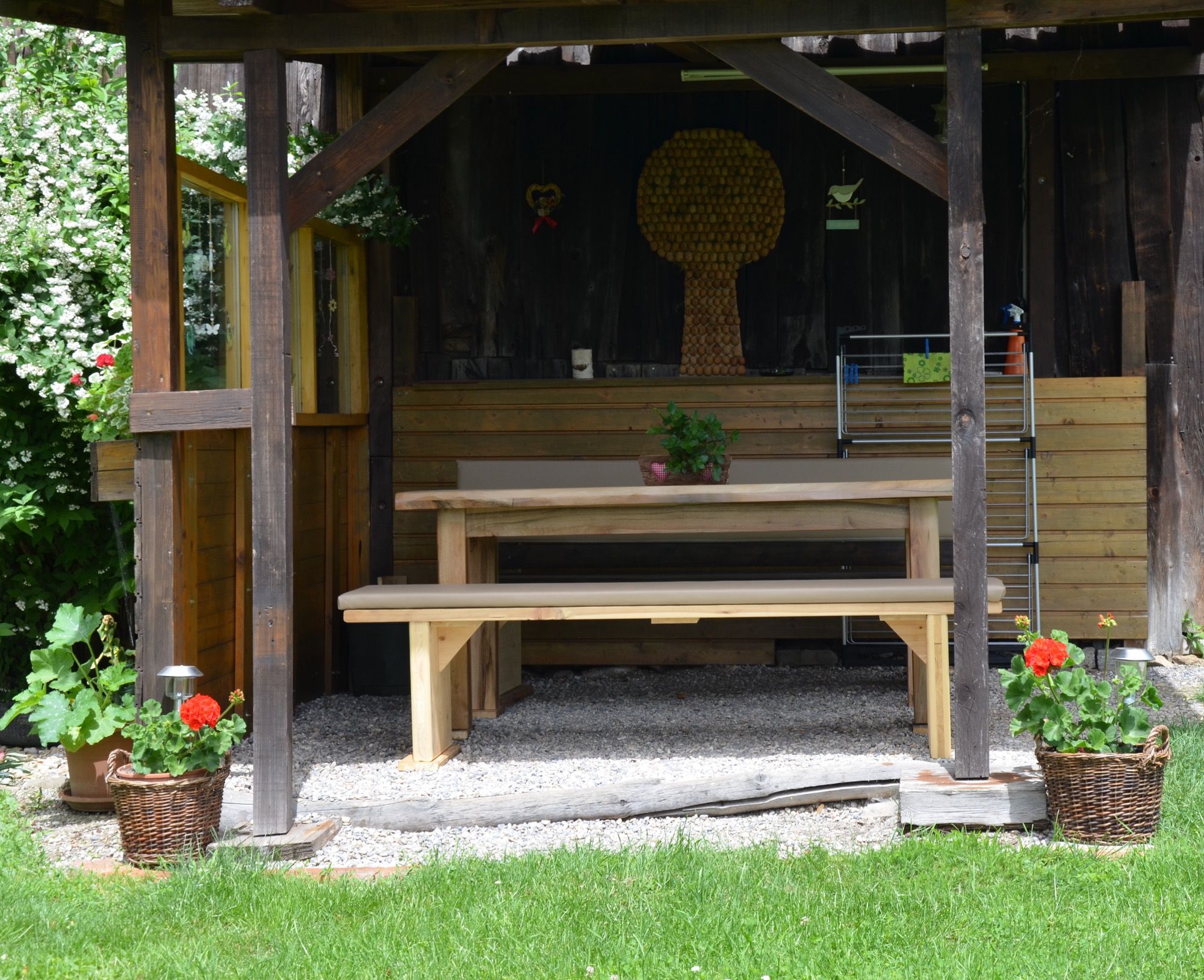 Egy rusztikus pavilon fa asztallal és padokkal, körülötte növényekkel és kosaras virágokkal.