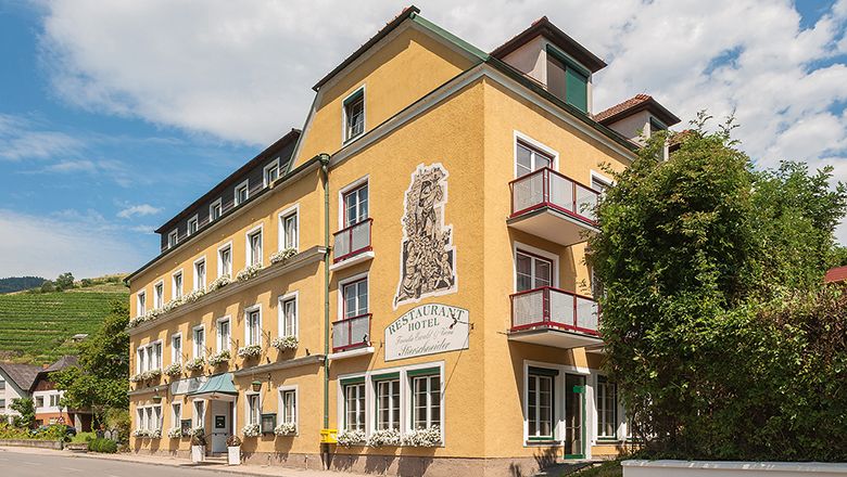 Sárga szállodaépület erkélyekkel és növényekkel, kék ég a háttérben.