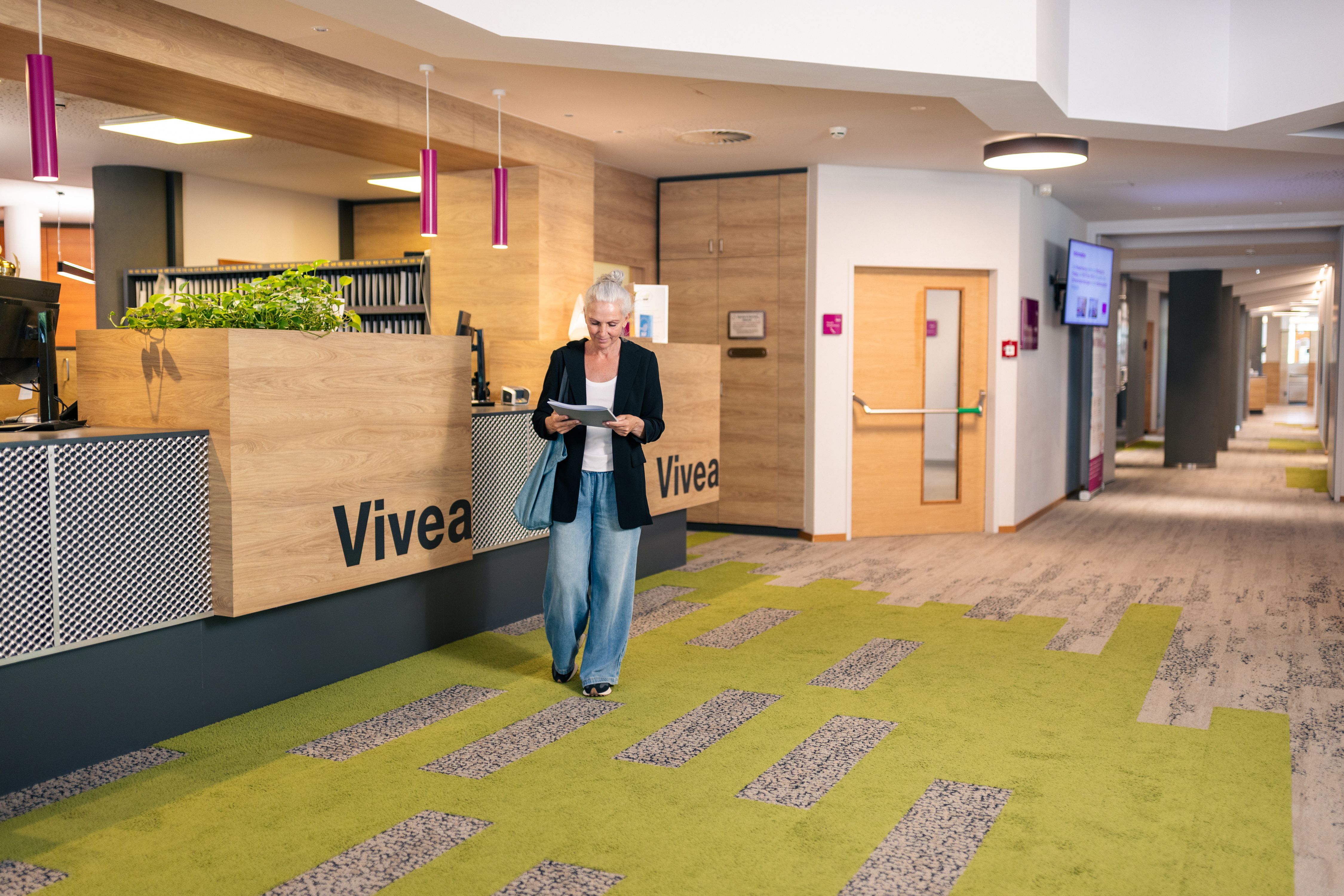Egy nő áll a Bad Schönau-i Vivea 4* szálloda előcsarnokában, és egy dokumentumot olvas. A recepció modern stílusban van berendezve: faelemekkel, növényekkel és egy szürke-zöld szőnyeggel.