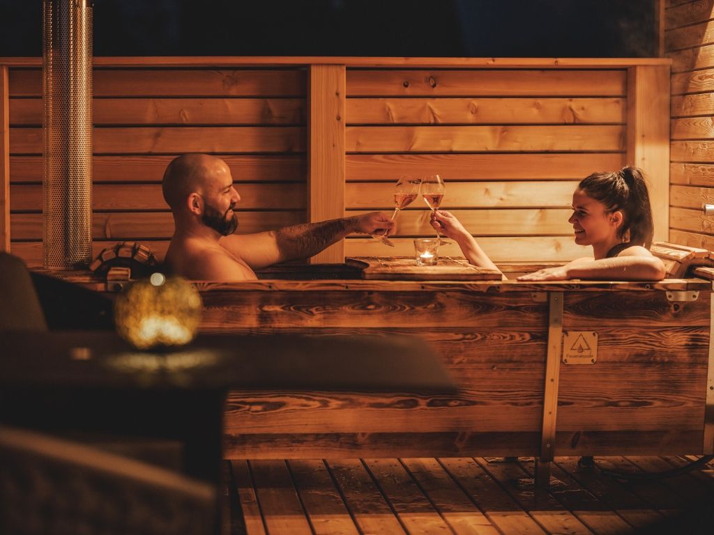 Két ember egy fából készült kádban a szabadban, borospoharakkal koccintva.
