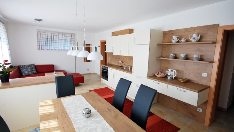 Modern nappali &eacute;s &eacute;tkező nyitott konyh&aacute;val, piros kanap&eacute;val, fa asztallal &eacute;s feh&eacute;r f&uuml;ggől&aacute;mp&aacute;kkal.