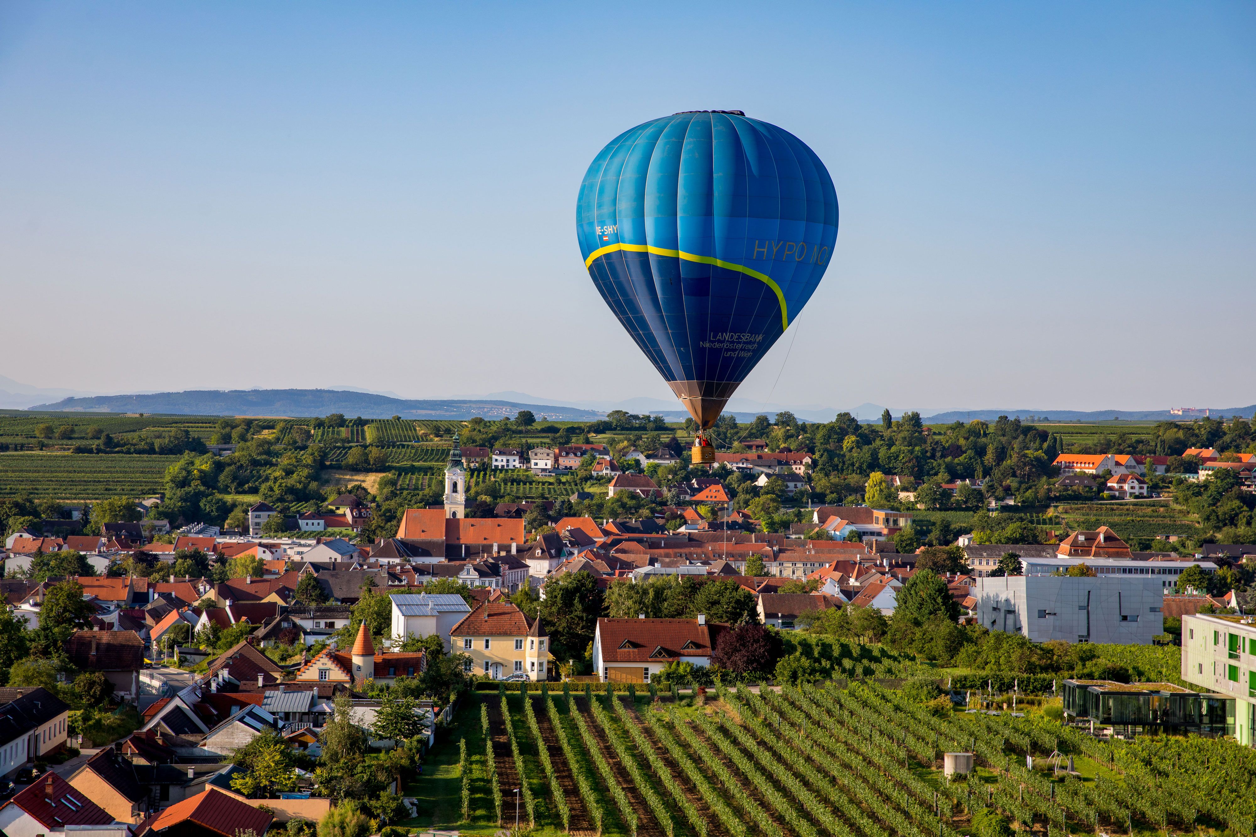 Hőlégballon egy szőlőskertekkel és házakkal tarkított városkép felett.
