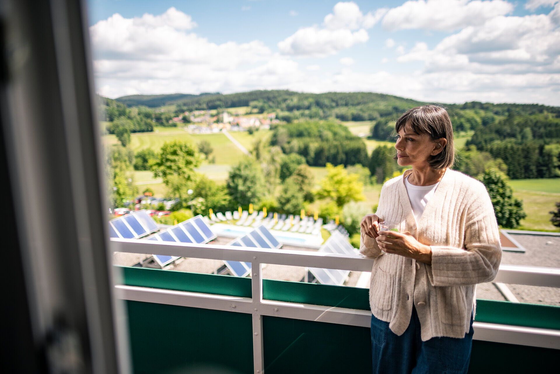 Egy személy egy csészével a kezében a Waldviertel régióban található Moorheilbad Harbach Egészségügyi és Rehabilitációs Központ erkélyén, kilátással a zöld dombvidékre.
