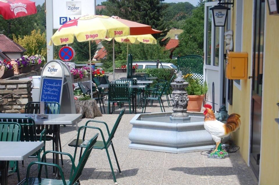 Vendégkert asztalokkal, székekkel, napernyőkkel és szökőkúttal. A szökőkút mellett egy kakas áll.