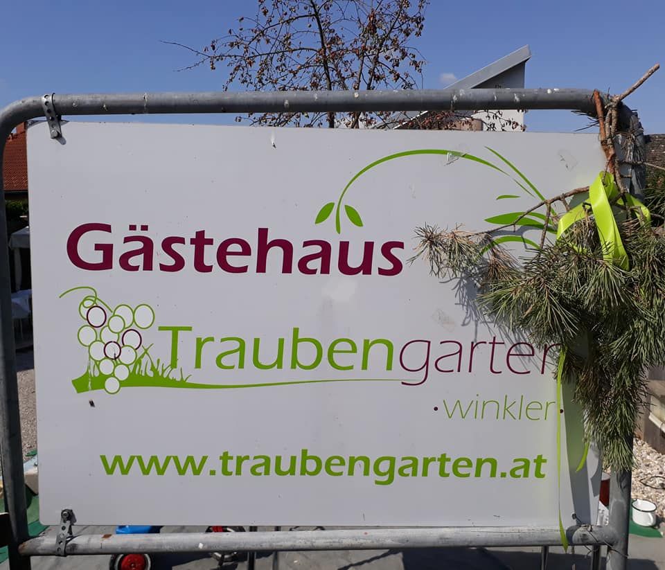 "Gästehaus Traubengarten Winkler" feliratú, ágakkal díszített tábla a "Gästehaus Traubengarten Winkler" felirattal és a weboldal címével.