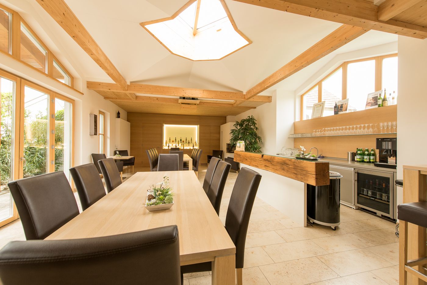 Belső nézet egy modern kóstolóteremről, fa asztalokkal és székekkel, nagy ablakokkal és egy bárpult palackozott borokkal.