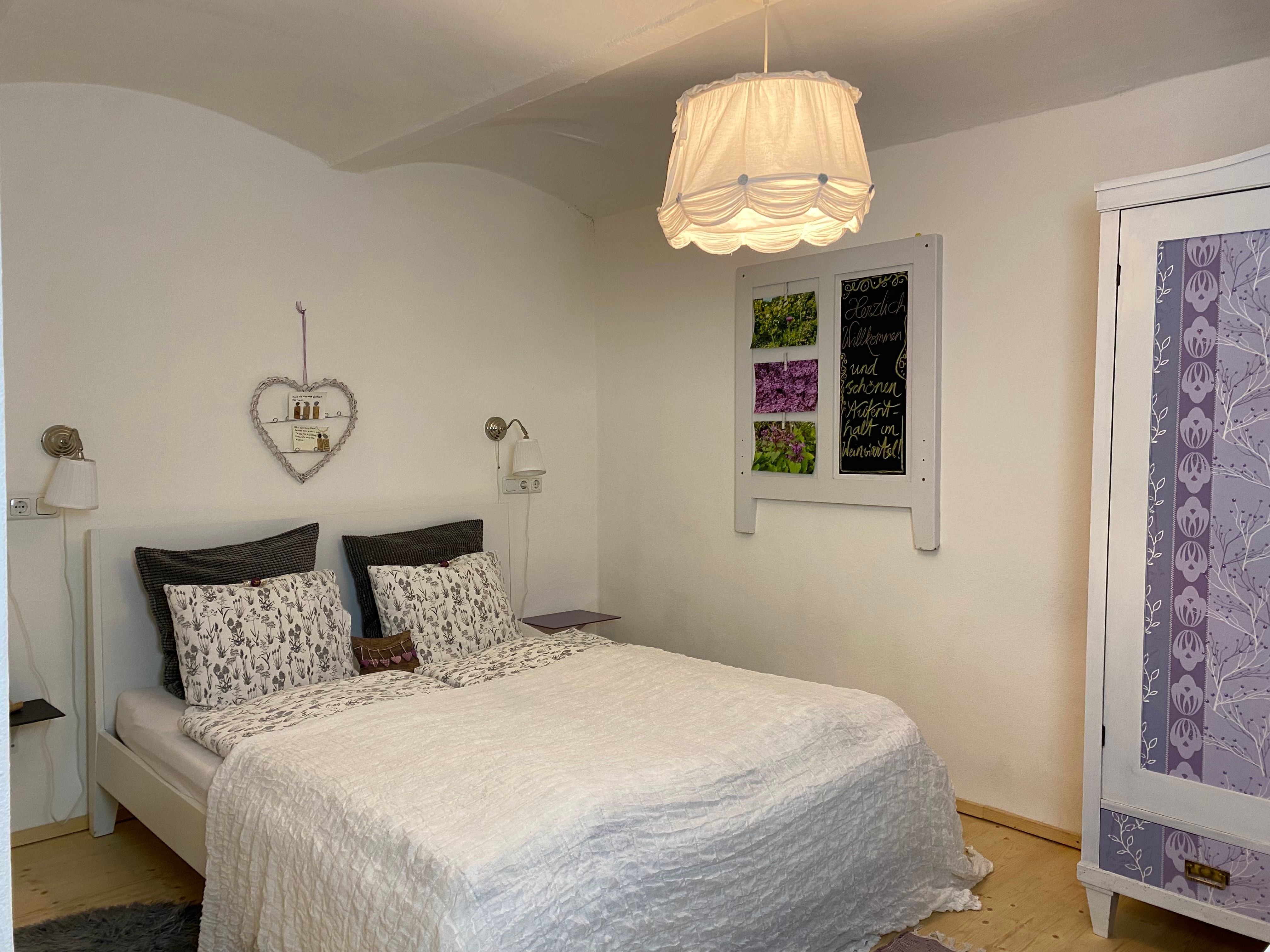 Hangulatos kétágyas szoba fehér ággyal, dekoratív szívpolccal és lila szekrénnyel.