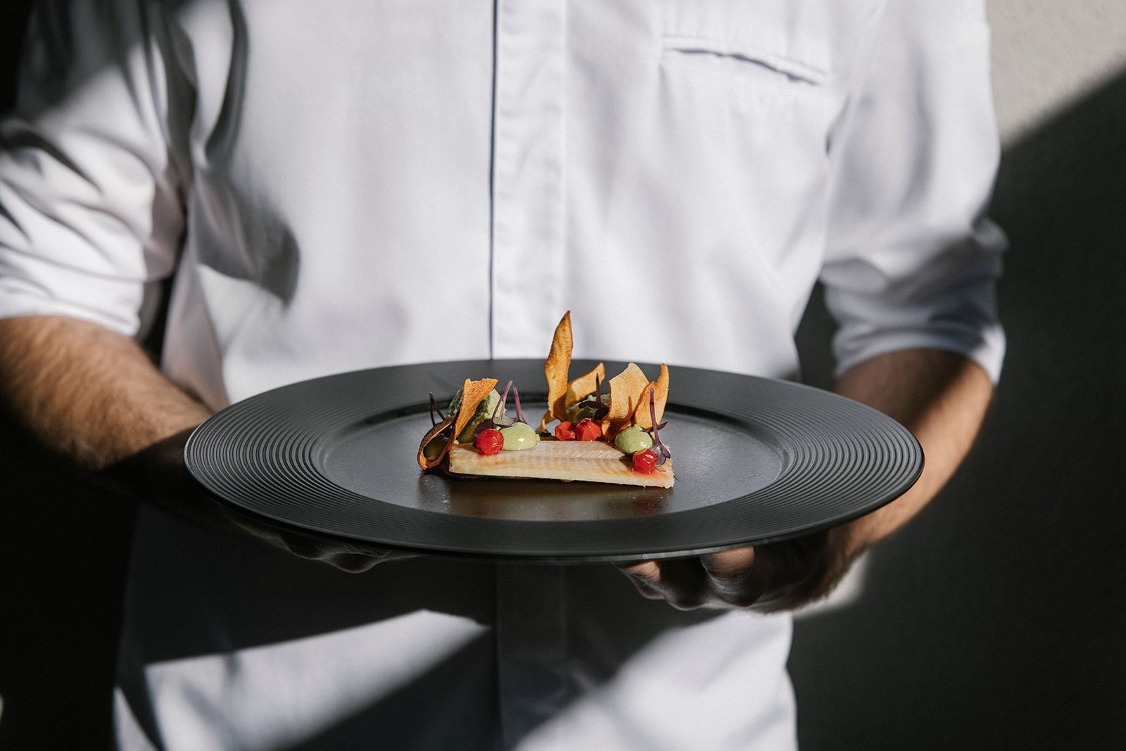 Egy fehér inges személy tányért tart egy tányérral, amelyen egy bonyolultan elkészített ínyenc étel van.