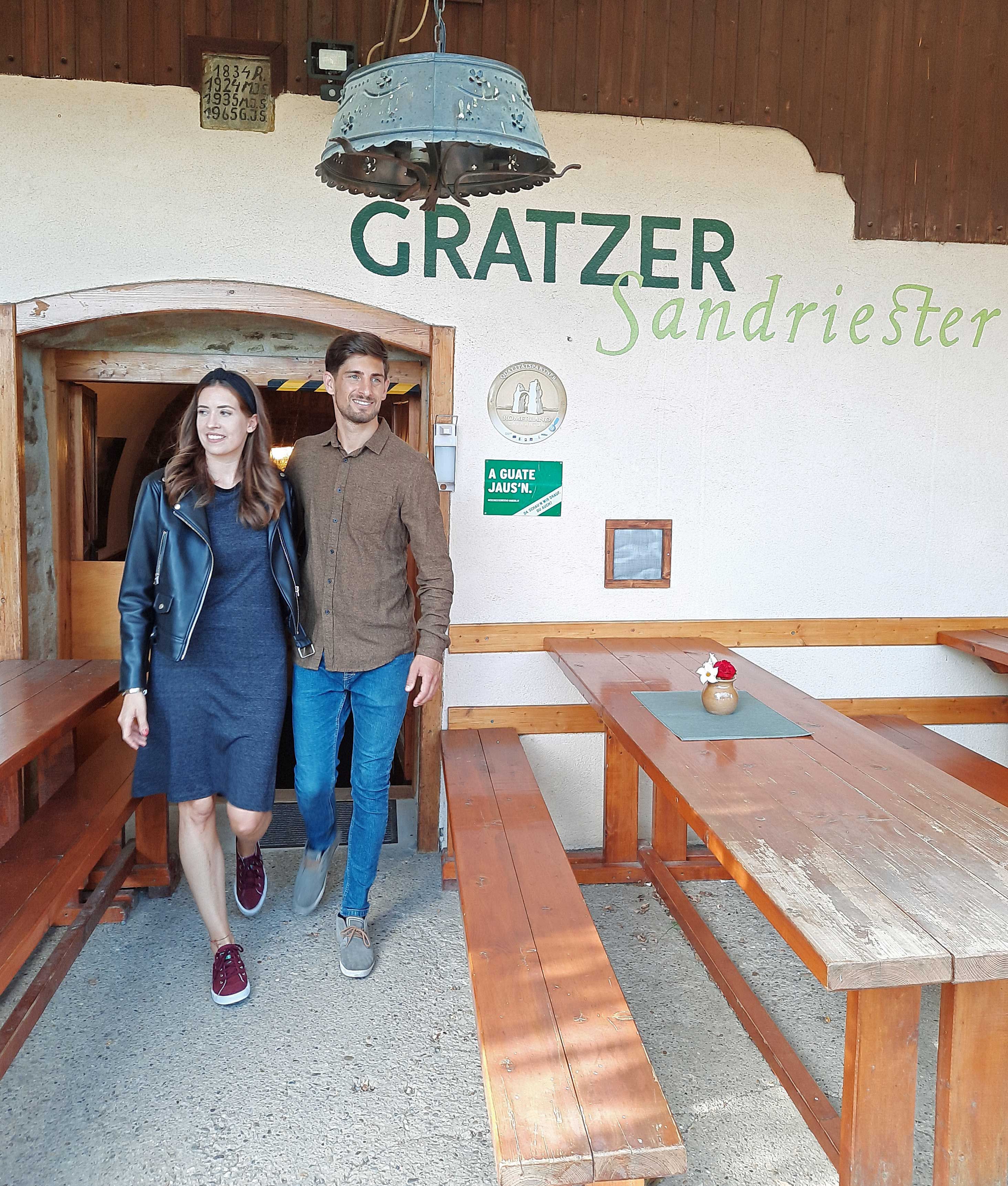 Egy férfi és egy nő elhagyja a Gratzer-Sandriester nevű hagyományos borozót. Mosolyogva sétálnak el a fapadok mellett.