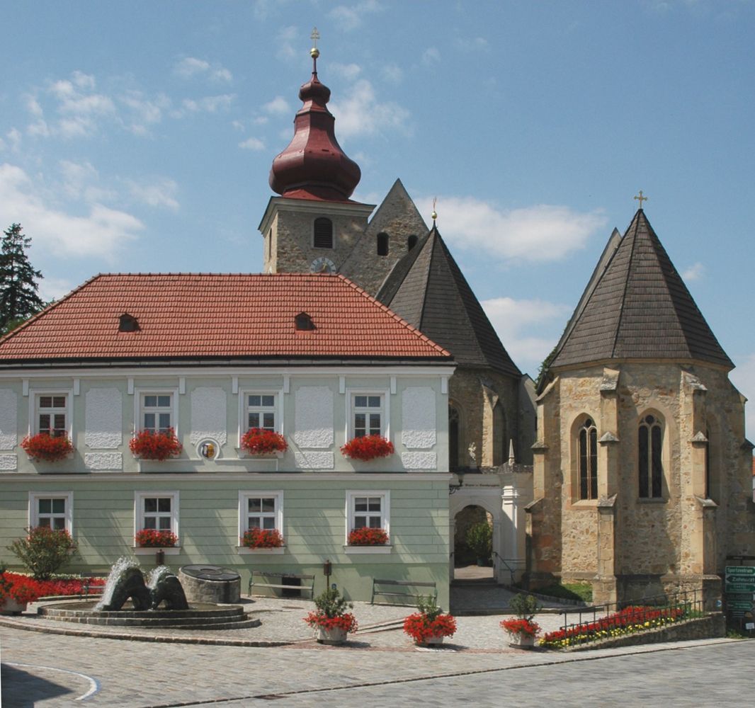 Történelmi épület vörös tetővel és templommal a háttérben a Maria-Anzbach piactéren.