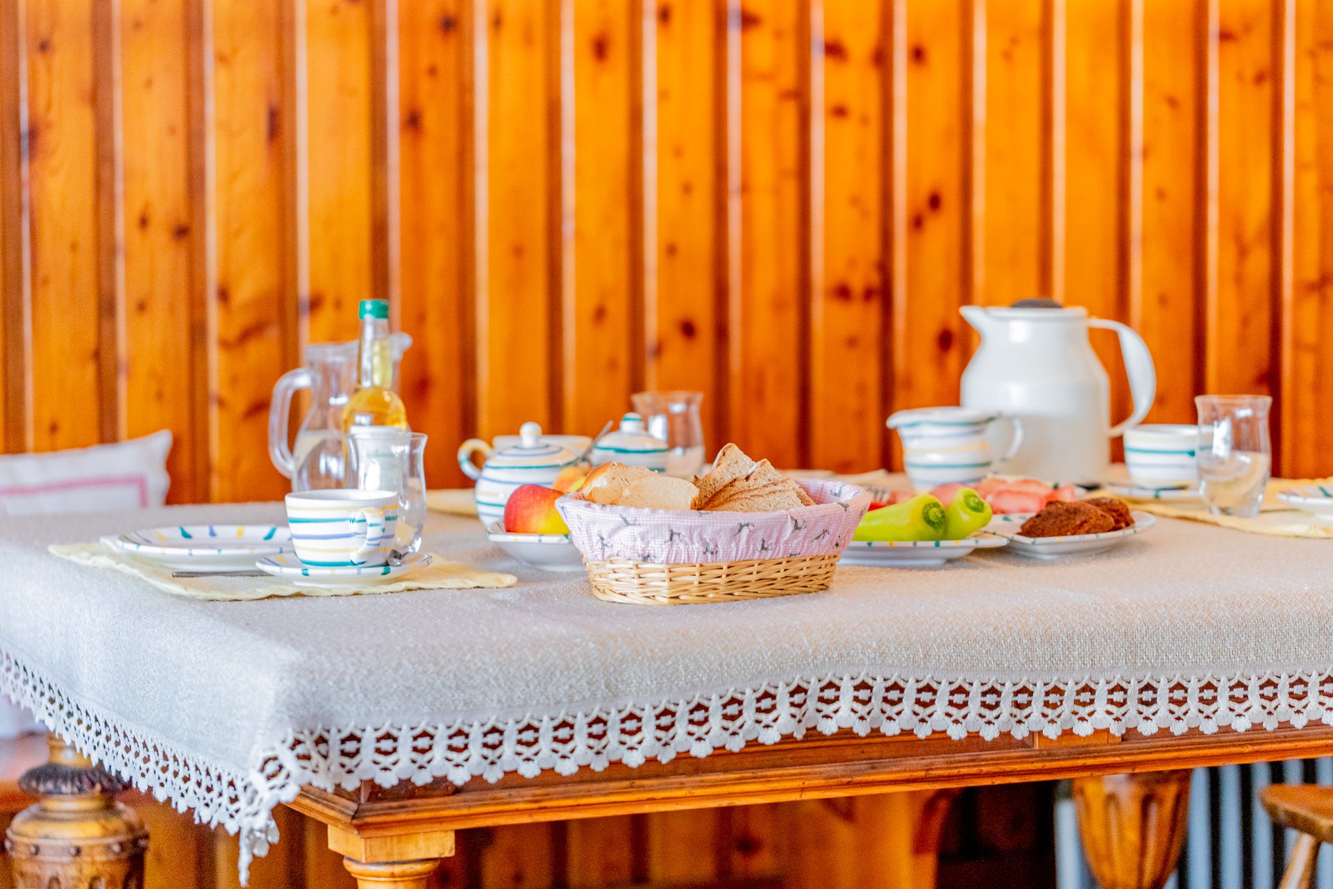 Reggelizőasztal edényekkel, kenyérrel, gyümölcsökkel és italokkal a faburkolat előtt.