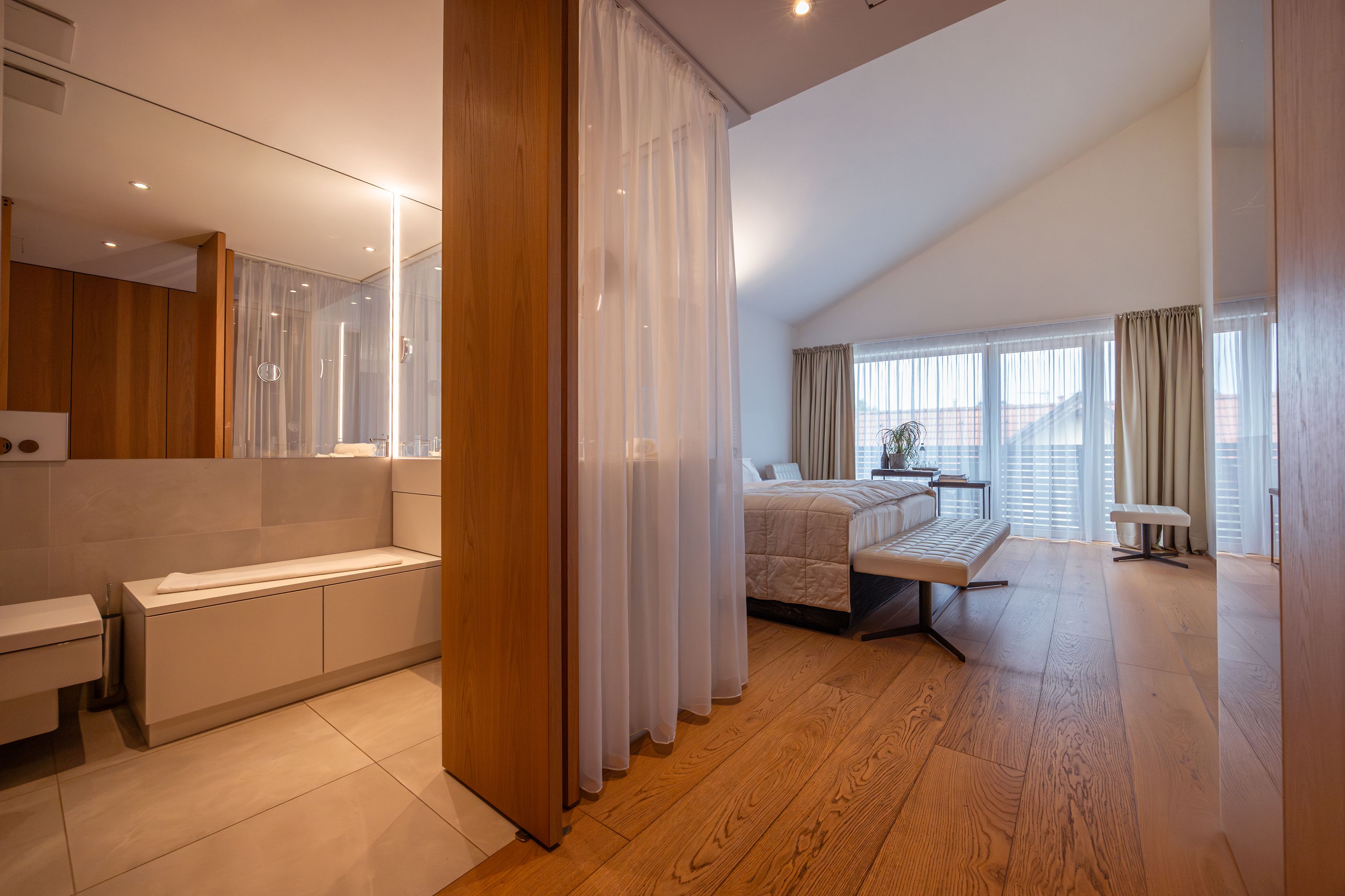 Modern szállodai szoba fapadlóval, nagy ággyal és tükrös fürdőszobával.