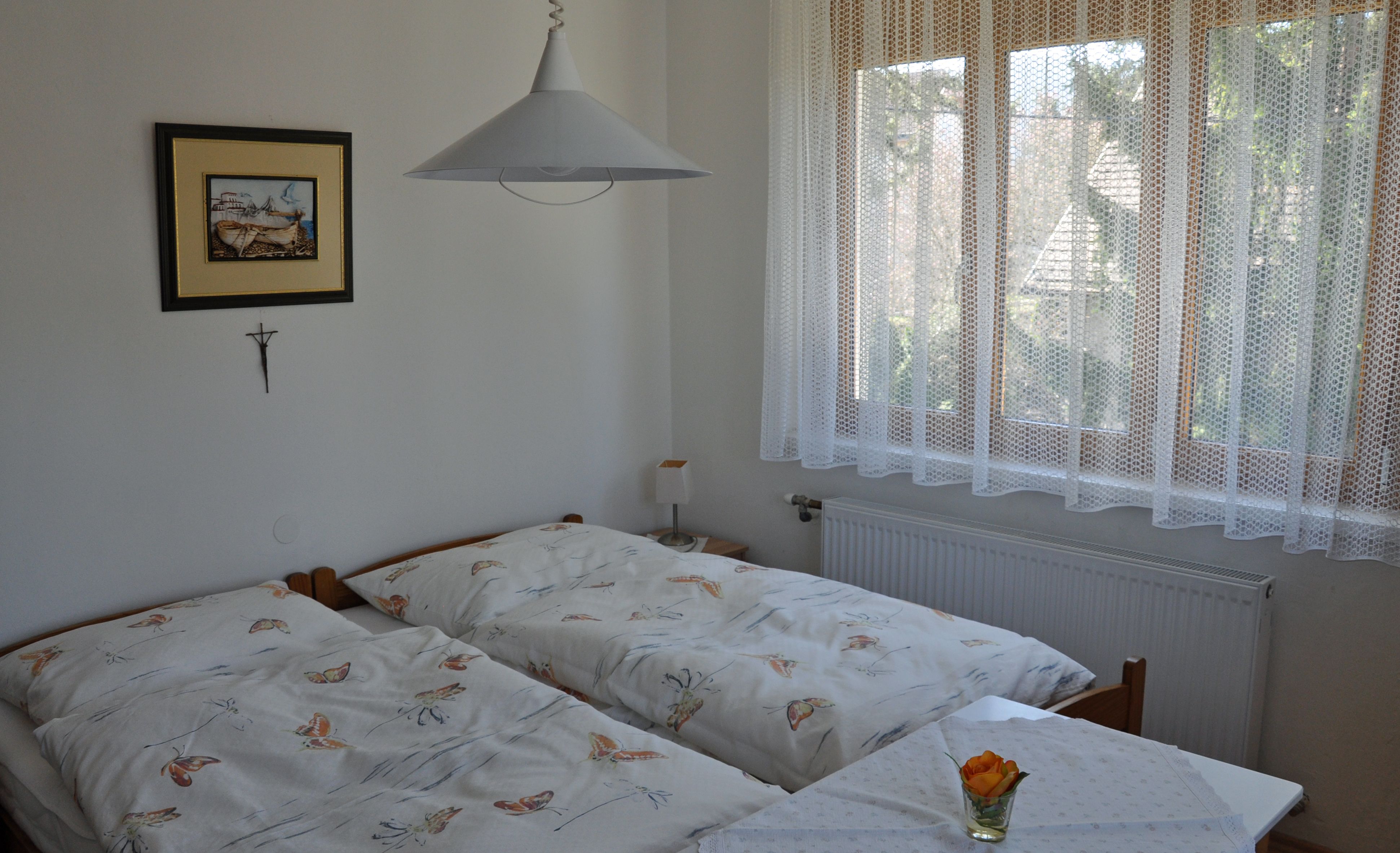 Hangulatos hálószoba két egyszemélyes ággyal, egy képpel a falon és egy csipkefüggönyös ablakkal.