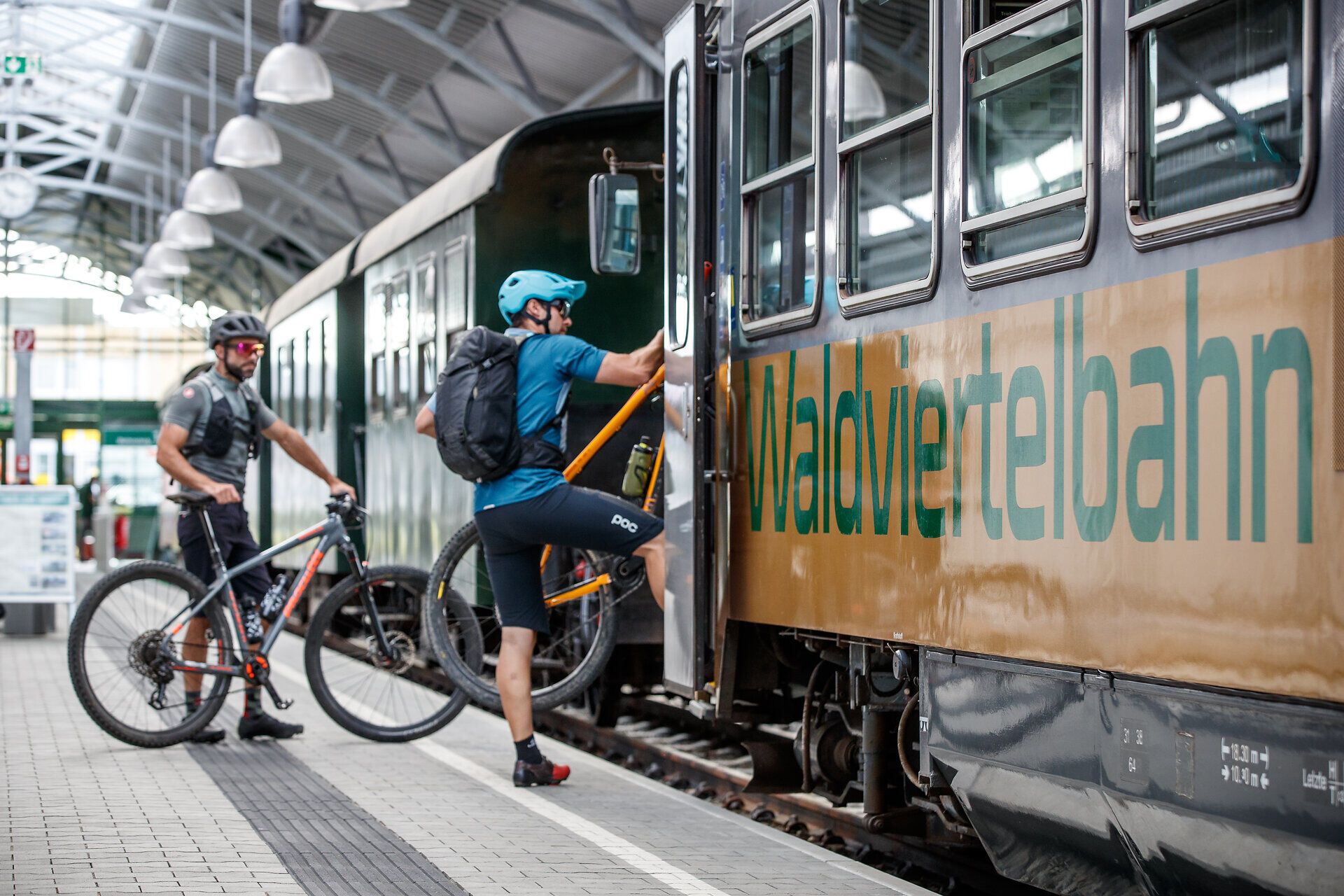 Ein Radfahrer steigt mit seinem Mountainbike in die wartende Bahn ein, während ein weiterer Radler auf dem Bahnsteig steht. Die Atmosphäre ist lebhaft und einladend, perfekt für Abenteuerlustige, die die malerische Landschaft des Waldviertels erkunden möchten.