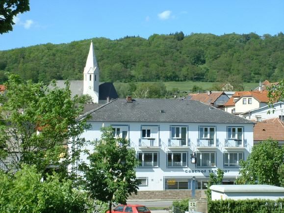 "Gasthof Schauer" feliratú kétszintes épület egy erdős domb és egy csúcsos tornyú templom előtt.