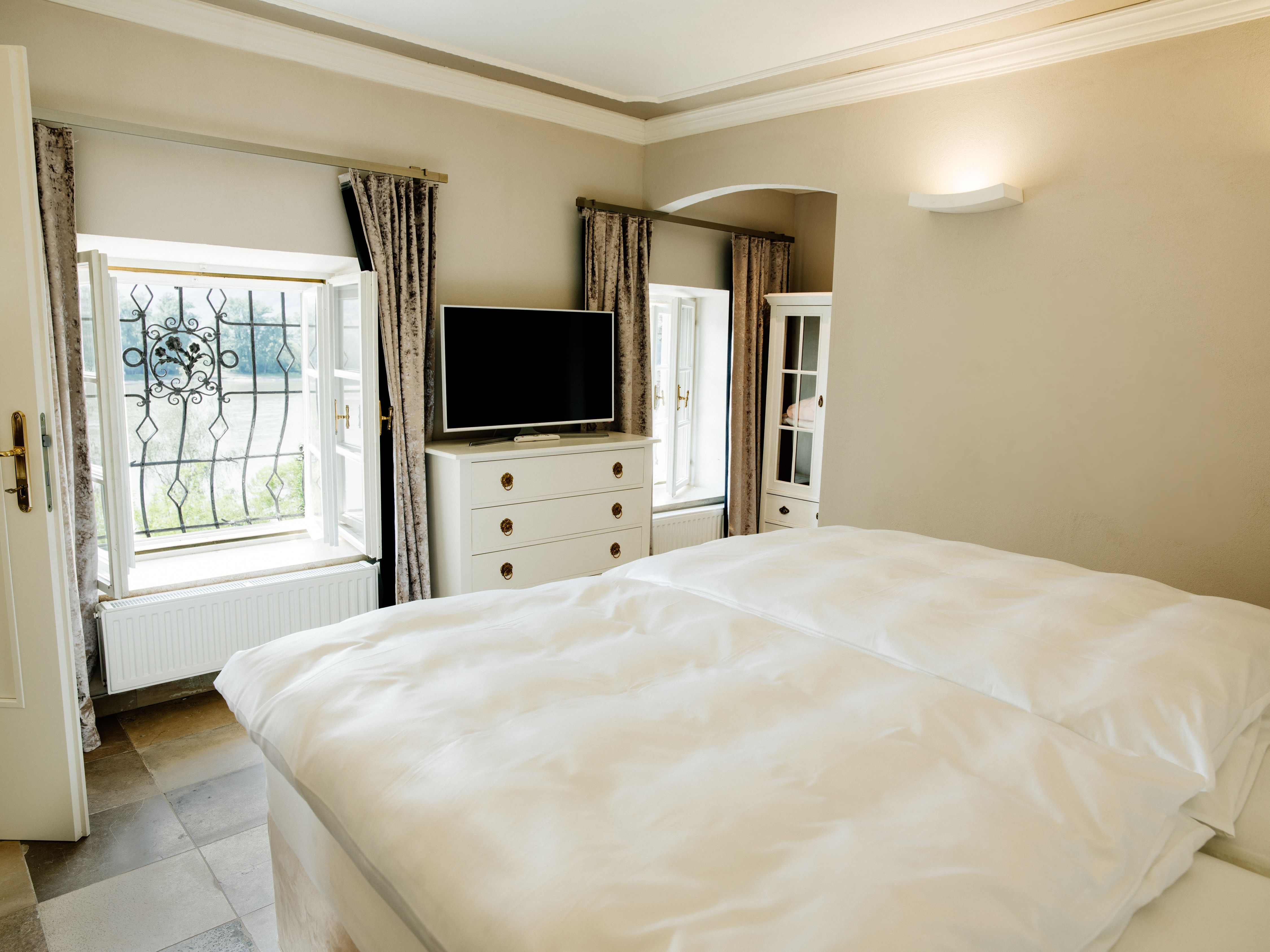 Világos hálószoba franciaággyal, TV-vel, komóddal és két függönyös ablakkal.