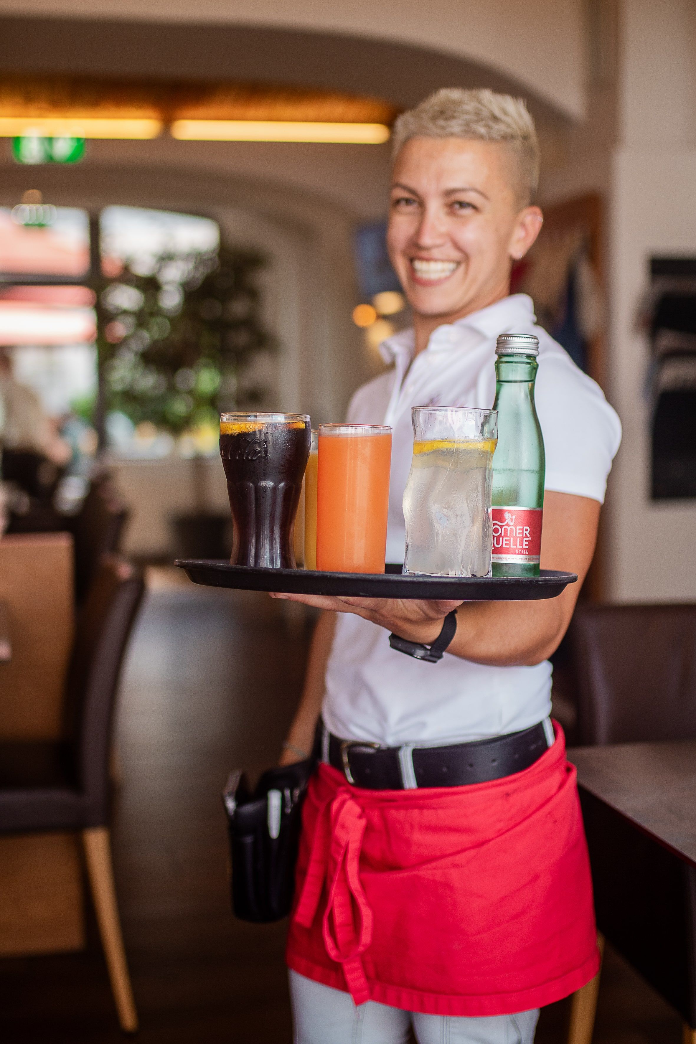 Egy mosolygós pincérnő egy étteremben egy tálcát visz különböző italokkal.