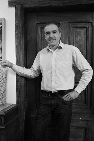 Egy ingben álló férfi áll egy faajtó előtt, és a kamerába mosolyog.