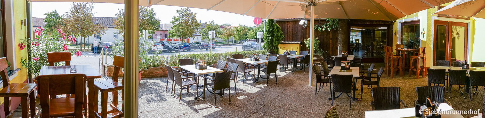 Napfényes vendégkert üres asztalokkal és székekkel, növényekkel és egy nagy napernyővel körülvéve.