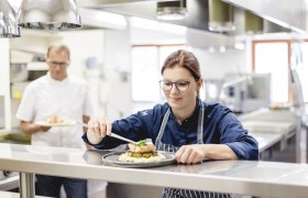 Egy séf egy étterem konyhájában egy ételt készít, miközben egy kollégája a háttérben egy másik ételt tart.