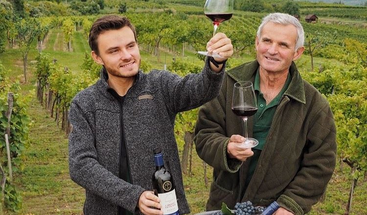K&eacute;t f&eacute;rfi egy szőlőben, kez&uuml;kben v&ouml;r&ouml;sboros poharakkal, mosolyogva. Egyik&uuml;k egy &uuml;veg bort tart a kez&eacute;ben; a h&aacute;tt&eacute;rben szőlőtők&eacute;k &eacute;s dombok l&aacute;that&oacute;k.