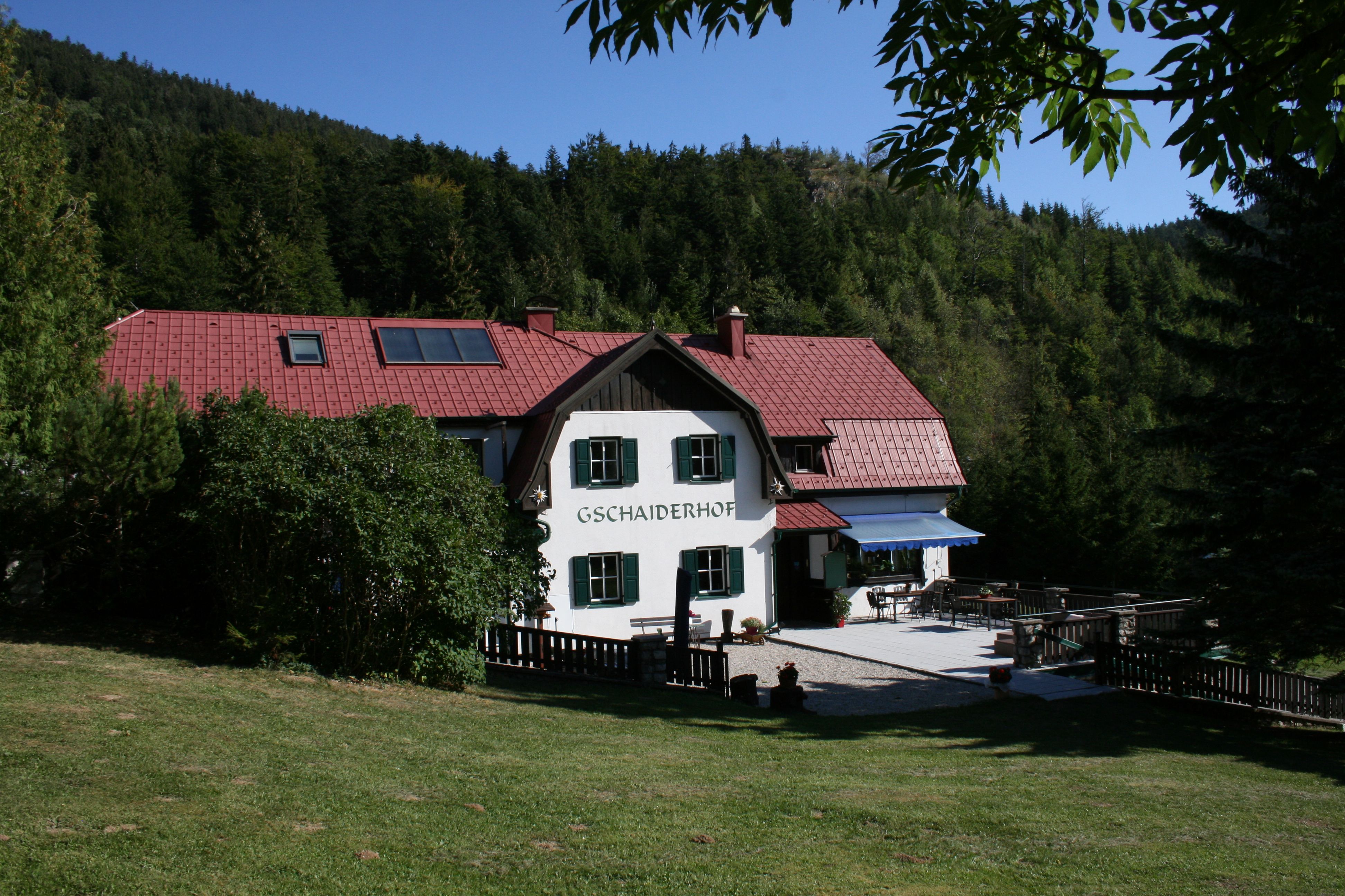 A "Gschaiderhof" feliratú, erdővel és rétekkel körülvett, vörös tetejű hagyományos ház.