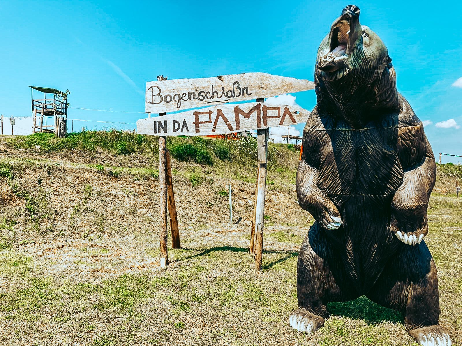 Egy nagy medvemodell a "Bogenschiaßn in da Pampa" feliratú tábla mellett egy réten.