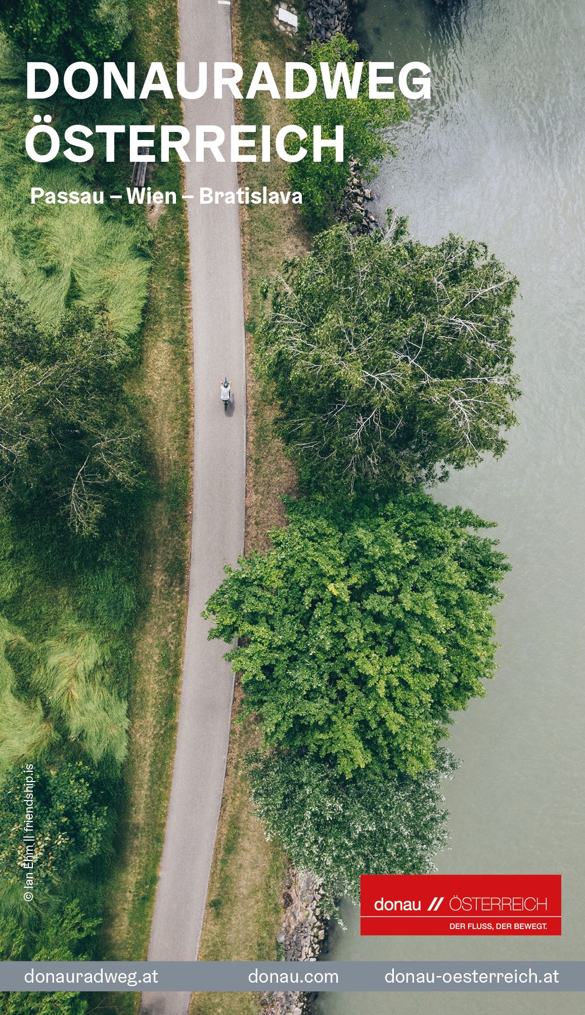 Eine Person ist mit einem Fahrrad auf dem Donauradweg unterwegs. Das Bild ist aus der Vogelperspektive fotografiert, man sieht links den Donauradweg umgeben von Bäumen und Wiese und rechts einen Teil der Donau. 