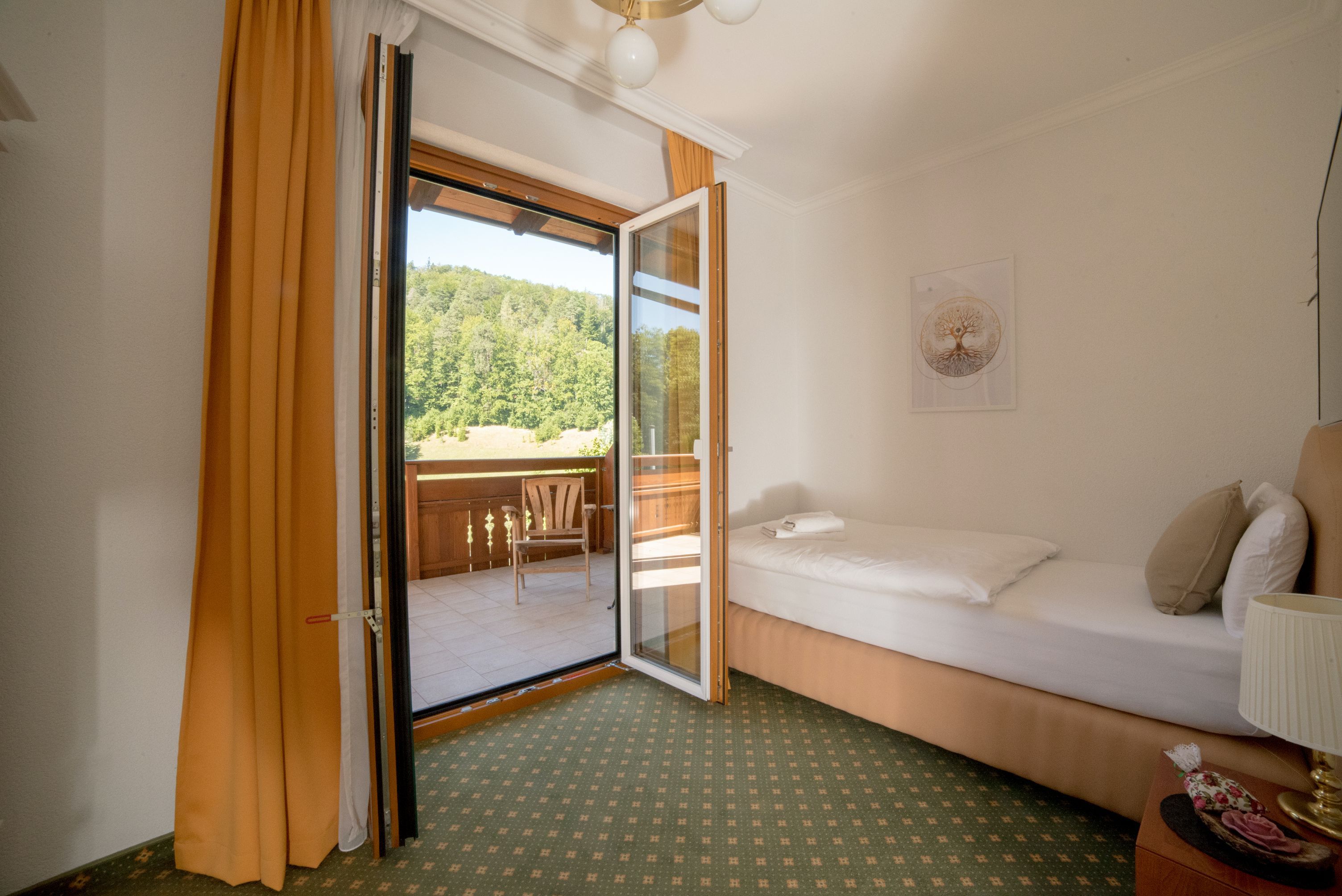 Egyágyas szoba erkéllyel, egyszemélyes ágy, nyitott erkélyajtó, kilátás a zöld tájra.
