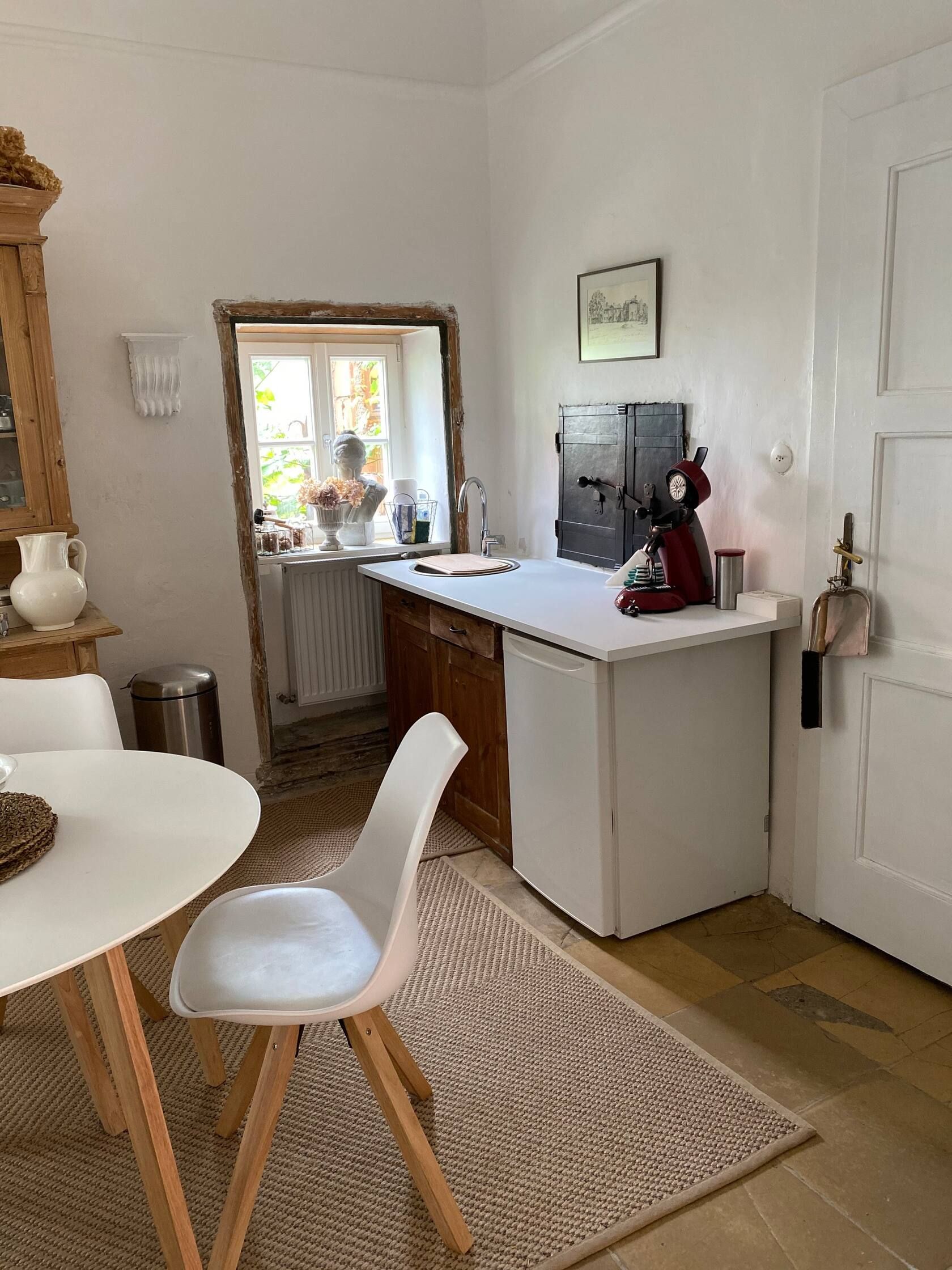 Konyha kis asztallal, székkel, mosogatóval és kávéfőzővel. Dekorációval ellátott ablakok és világos falak.