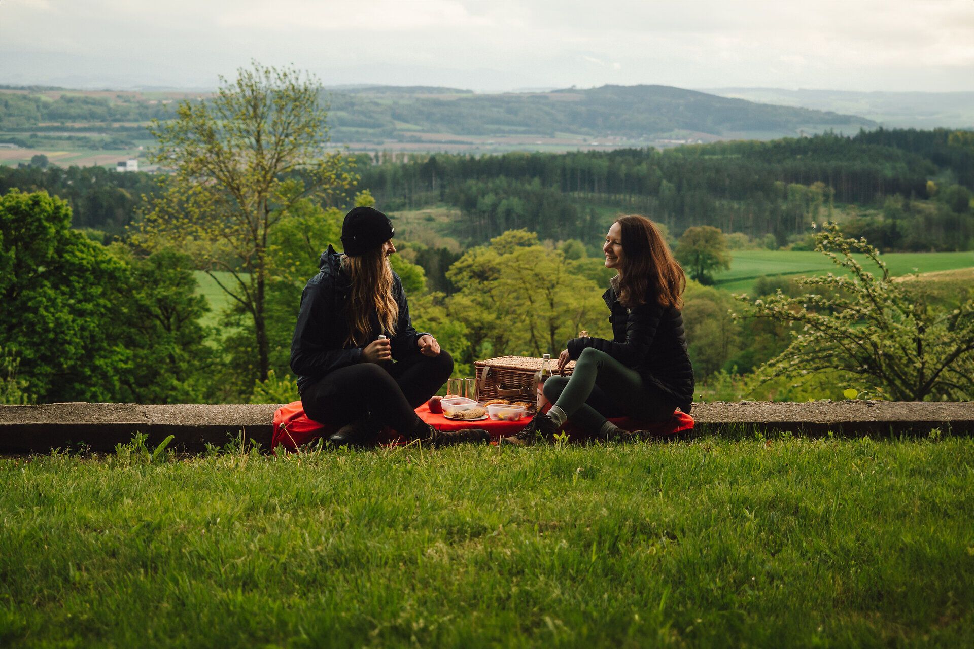 Két barát egy festői táj közepén, egy réten piknikezik. A buja zöld növényzettel és dombokkal körülvett táj békés hangulatot áraszt, amely elidőzésre hív.