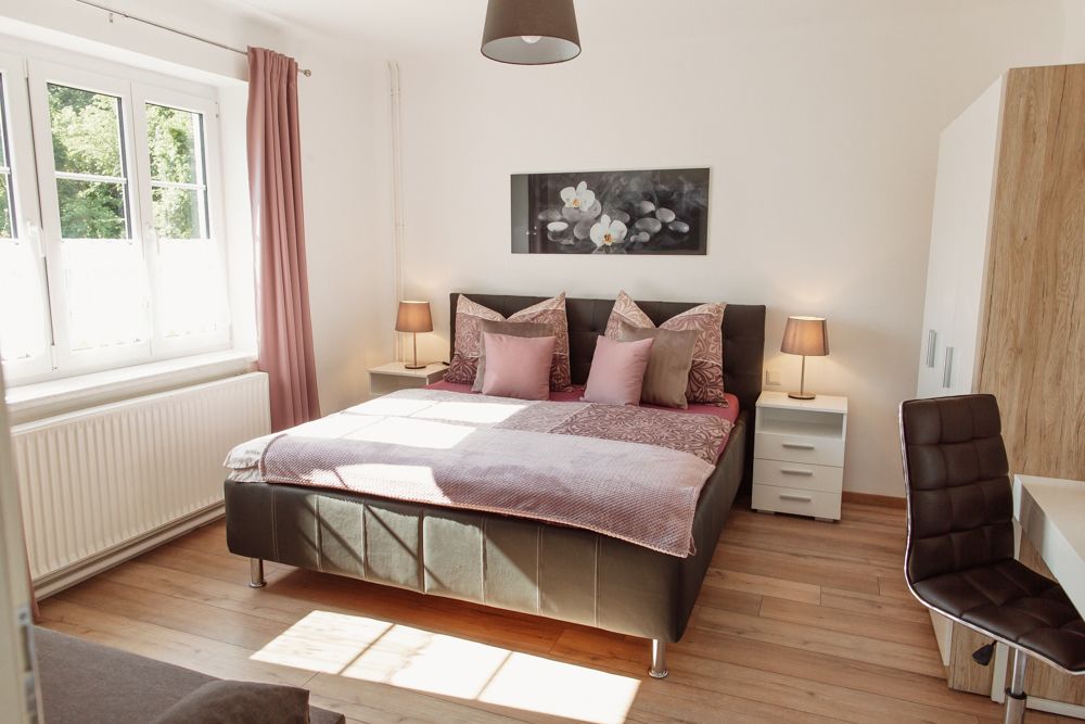 Világos hálószoba franciaággyal, két éjjeliszekrénnyel és rózsaszín függönyös ablakkal.