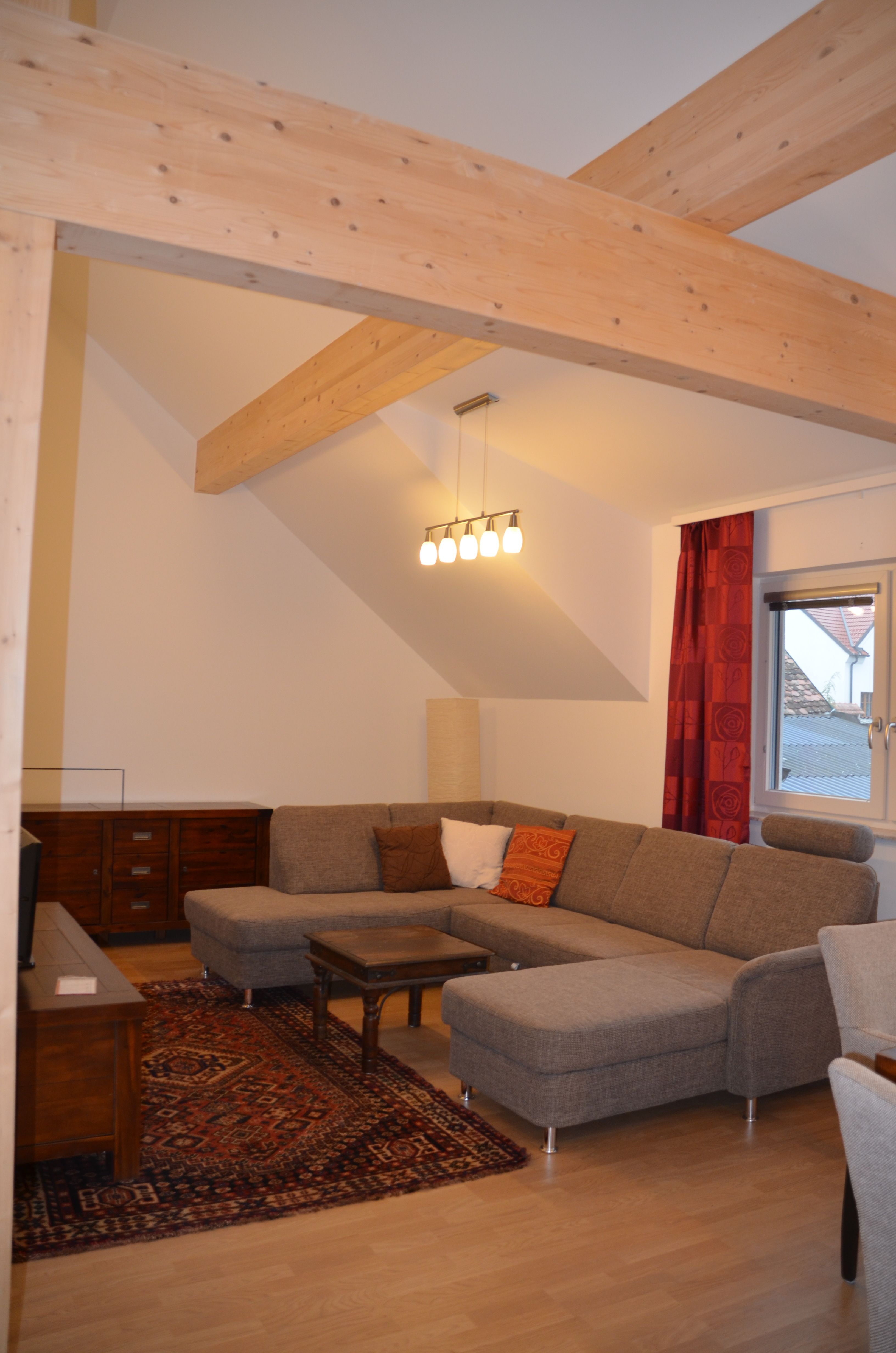 Modern nappali szürke sarokkanapéval, fagerendákkal, szőnyeggel és lámpával.