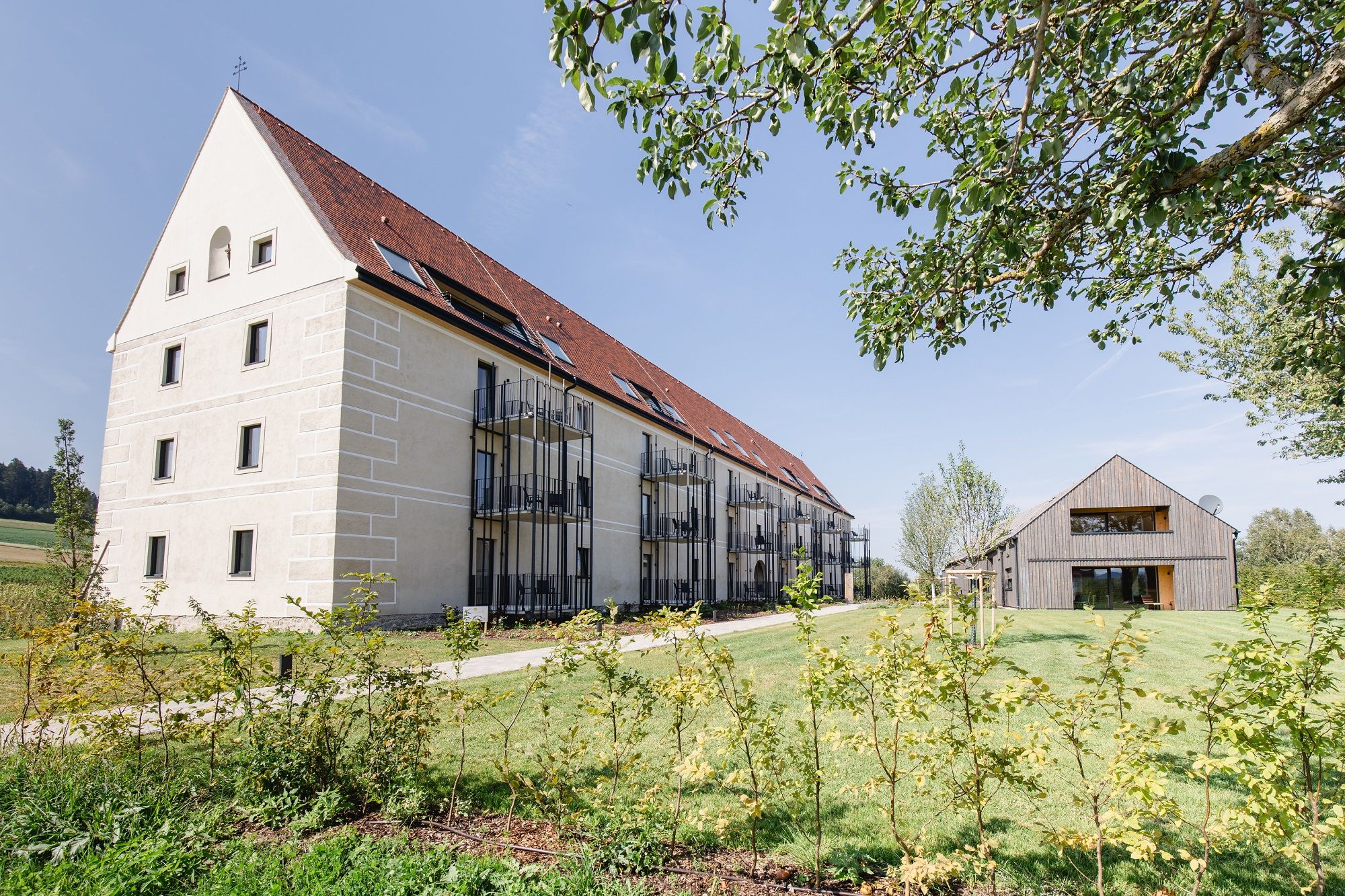 Egy nagy, modern, piros tetővel és erkélyekkel ellátott épület egy kisebb faház mellett, zöld telken.