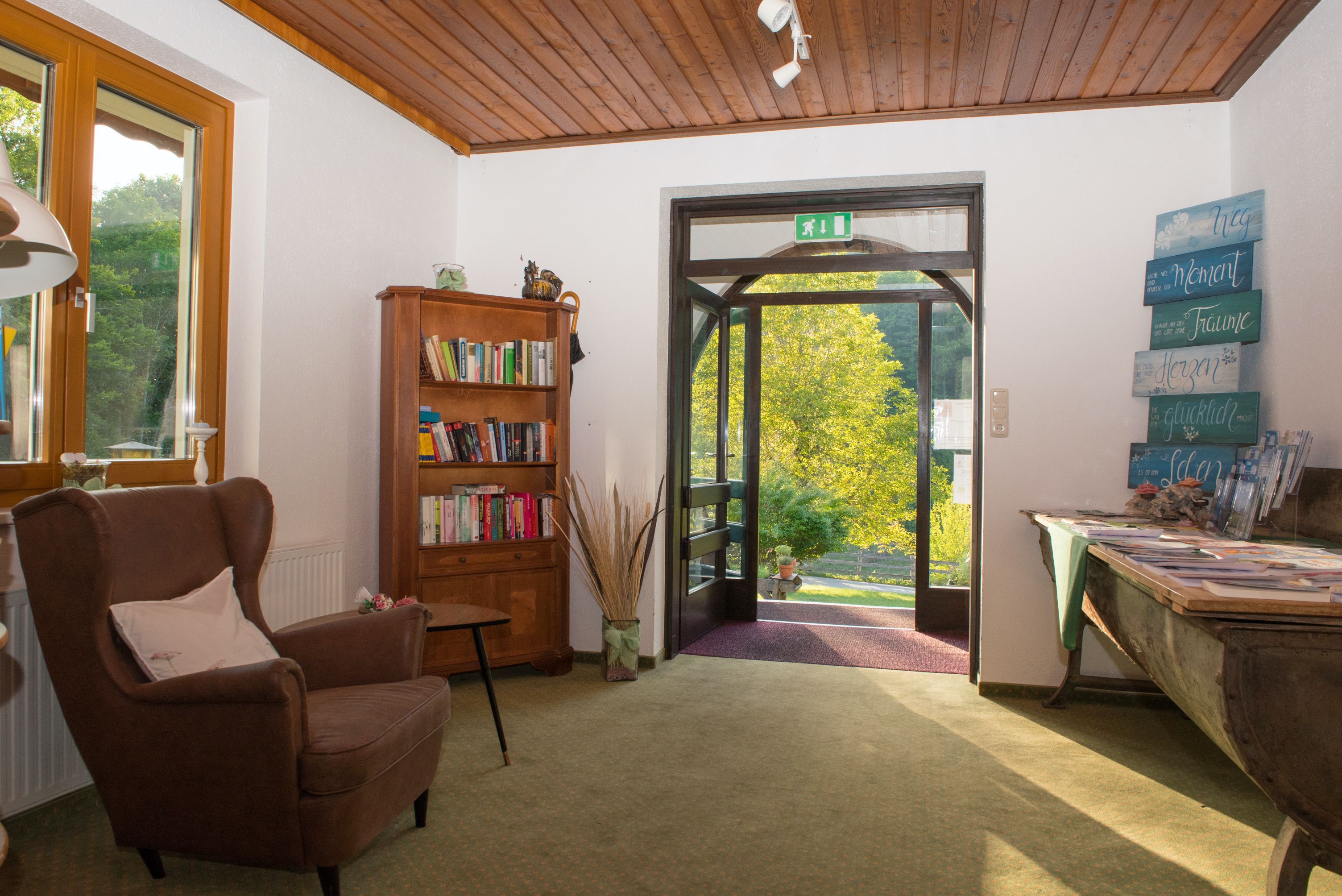 Bejárati rész karosszékkel, könyvespolccal és nyitott ajtóval, ahonnan kilátás nyílik a zöldövezetre.