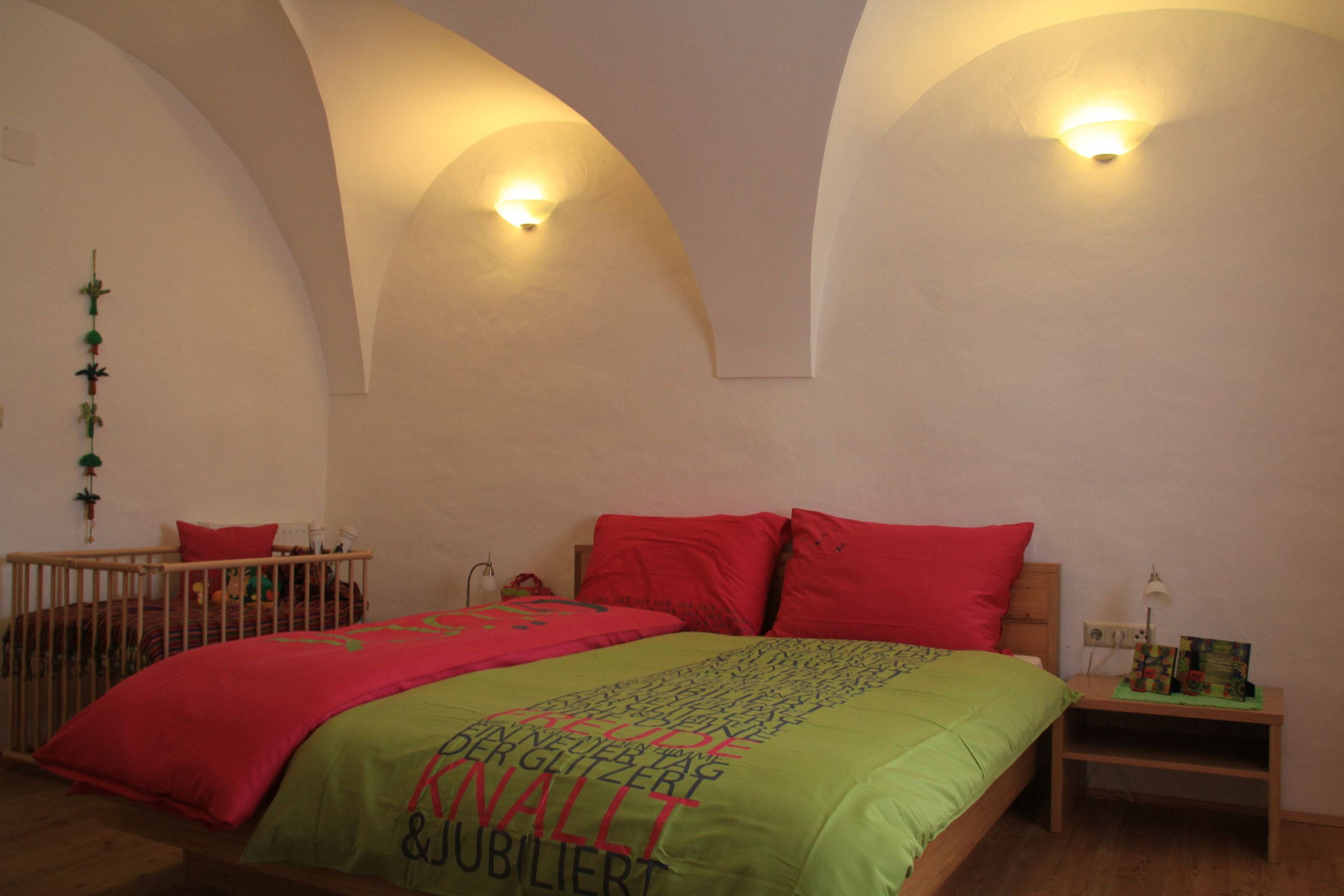 Hangulatos vendégszoba franciaággyal, színes paplanhuzatokkal és a háttérben egy babaágy. A falak fehérek és boltívesek.